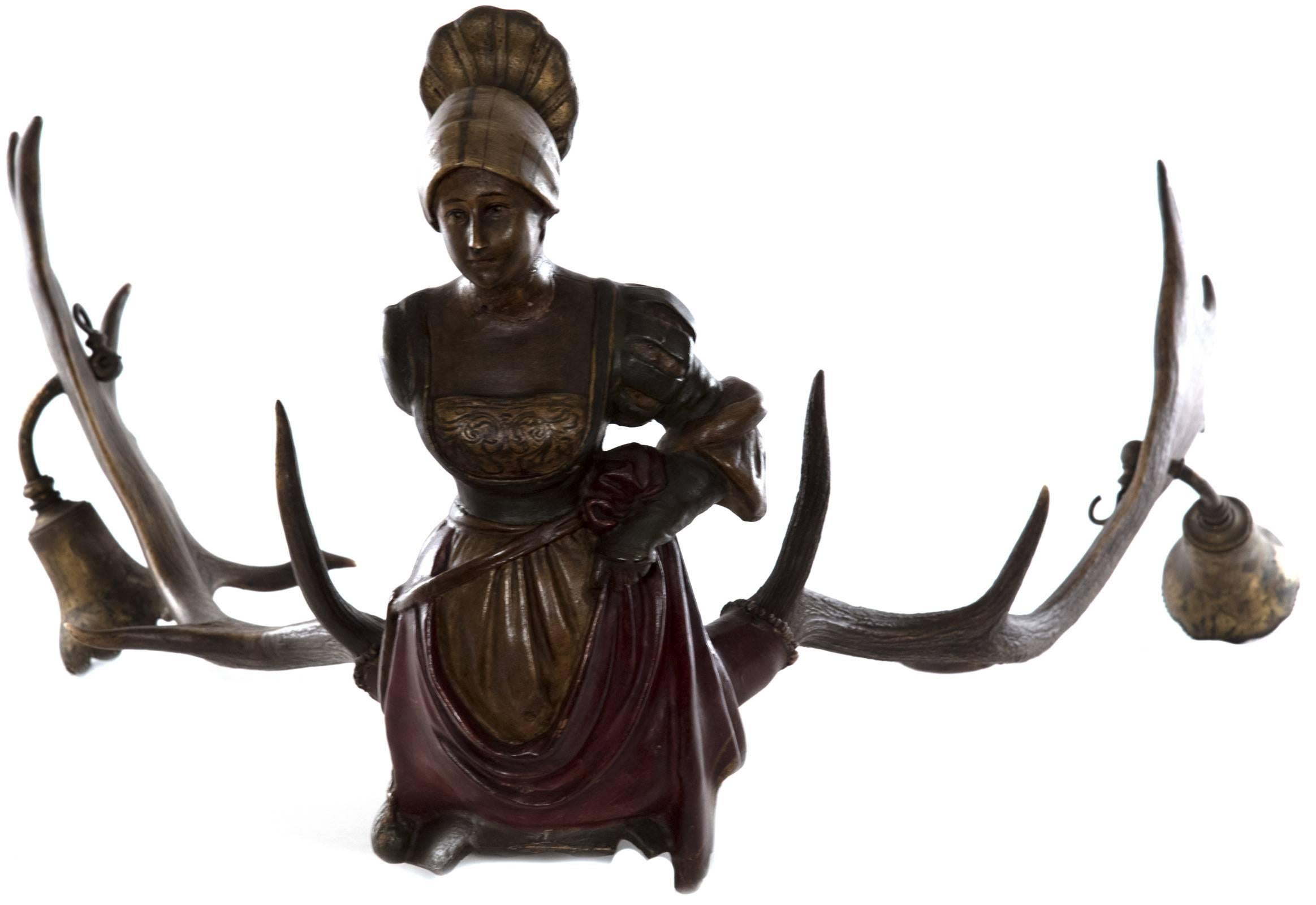 A Lu¨sterweibchen of female form in a richly colored dress and upturned head wrap, with a shield form on the base that depicts an idyllic image of two cherubs smelling roses on a garden bench. Two brass trumpet shaped light fixtures have been