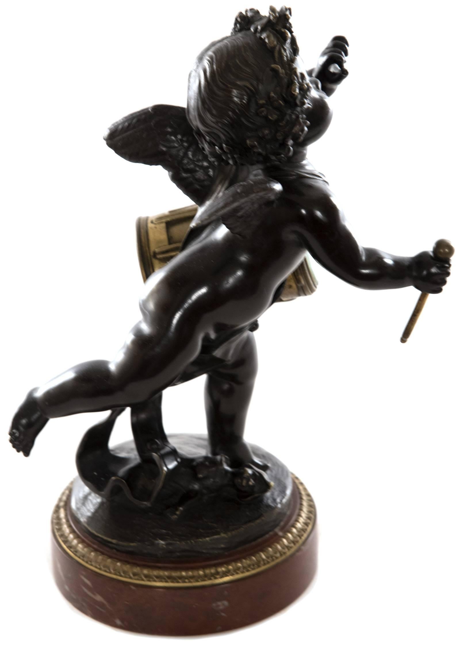 A French bronze desk clock in the form of a cherub holding a drumstick and drum, which encases the clock movements, on a round red marble base. The circular white enamel clock face is delicately decorated with floral swags hung by blue ribbons, with