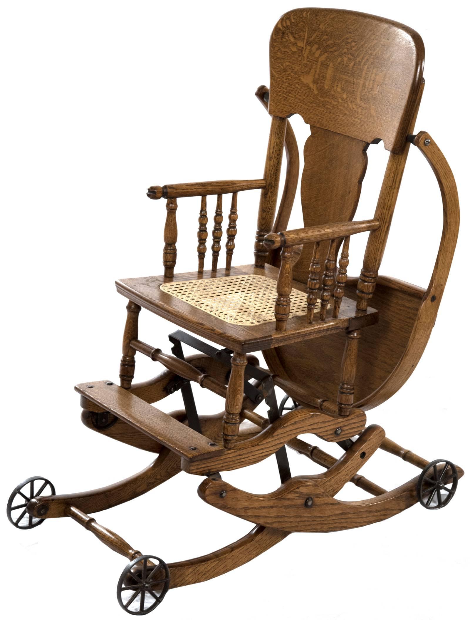 This solid golden oak baby highchair is composed of turned spindles and supports, a cane seat, a deep tray that lifts to move behind the seat, and sits on four metal wheels. The mechanics of the construction allow the highchair to convert to a