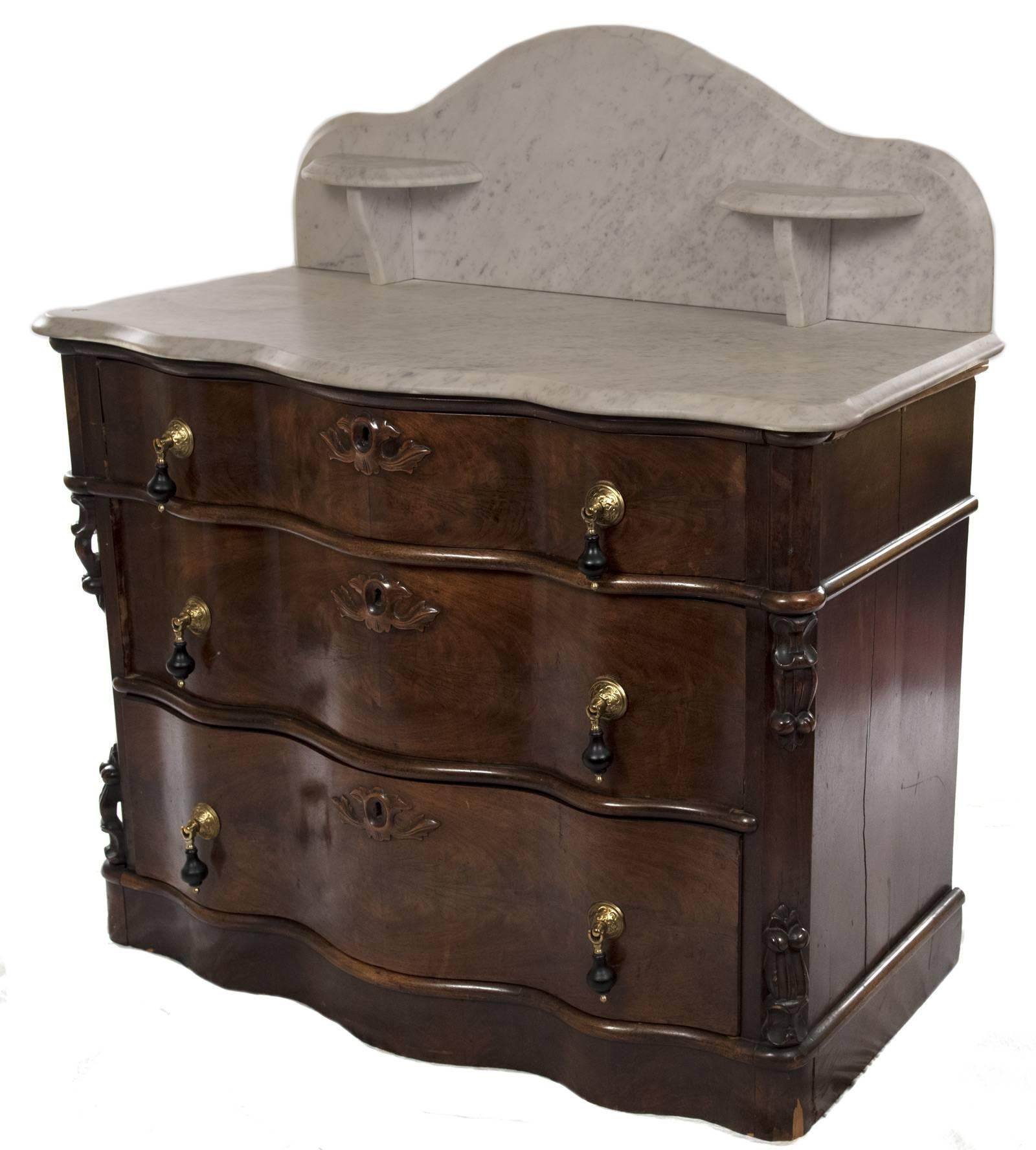 A marble backsplash and topped serpentine washstand with conforming walnut base with brass and ebonized drawer pulls and carved walnut keyhole design mounted on graduated drawers that are flanked with carved jambs and all sitting on a flat bottom.