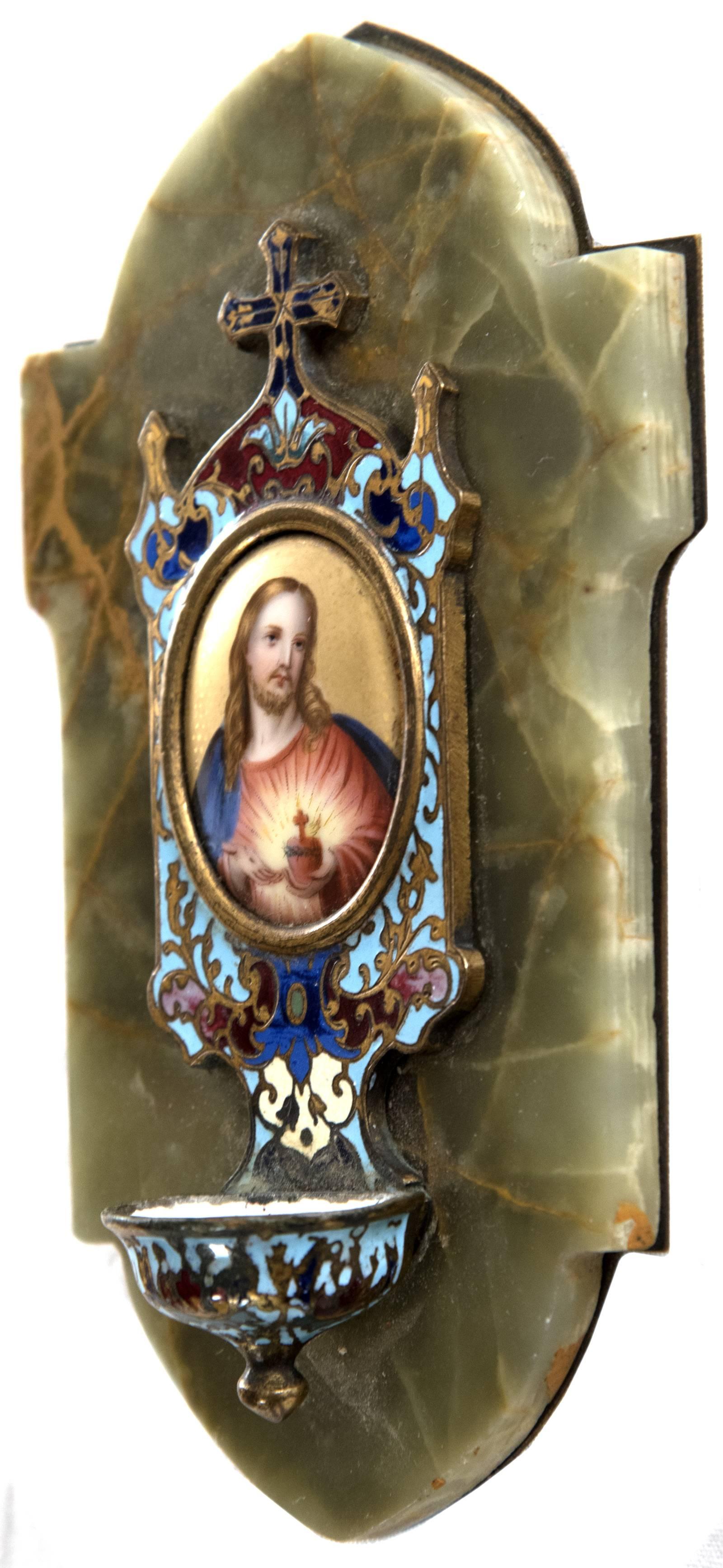 A hand-painted image of Jesus Christ on a porcelain oval is surrounded by a champleve enamel frame, with surmounting finals and centre cross, terminating in an attached water font, on green and gold-veined marble that is attached to a brass