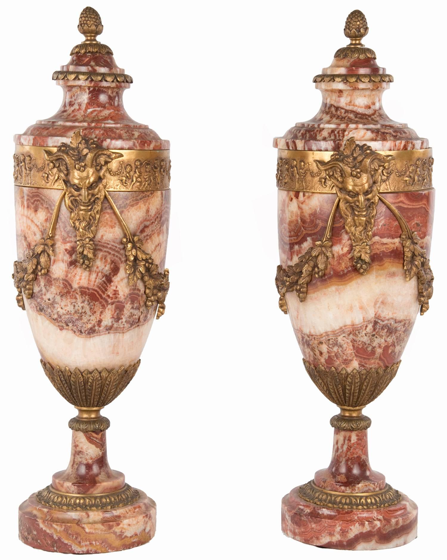 A pair of red and white breche marble urns of baluster form that are decorated with an ormolu band of dancing putti and satyr, or Bacchus, heads, from which hang a garland of grape bushels and leaves. Rings of gilt bronze acanthus leaves further