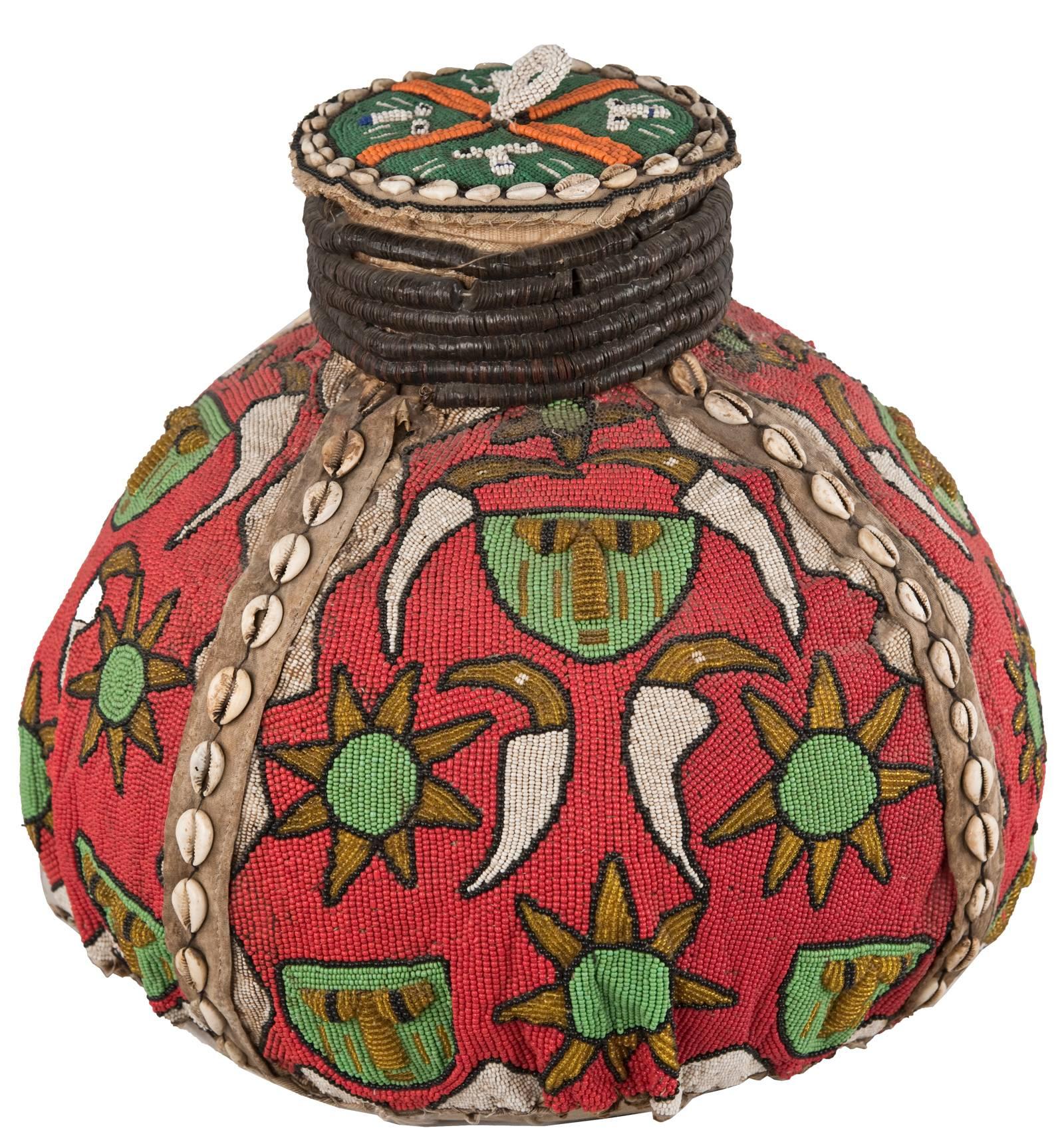 A beaded gourd with lid from the Yoruba people of Nigeria having a red ground decorated with stylized sun medallions and faces, and accented with bands of cowrie shells. Beads were a sign of wealth and status, and many Yoruba sacred and secular