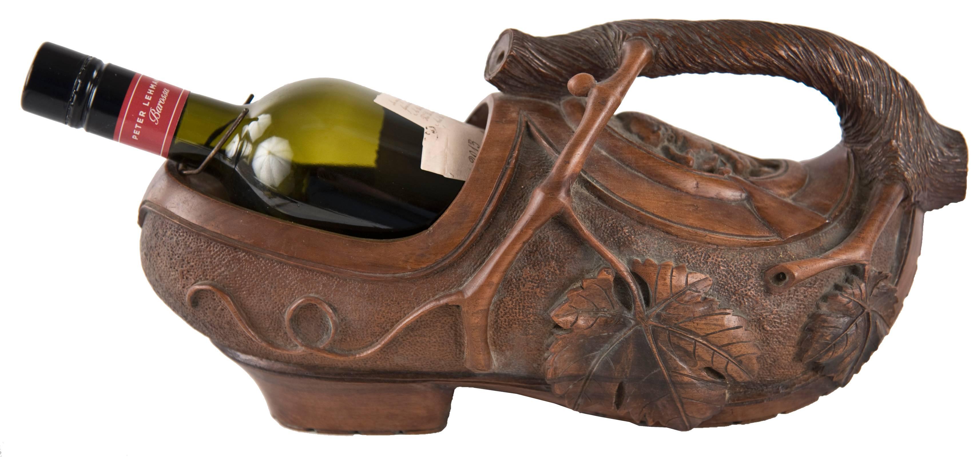 A carved wooden wine bottle holder in the form of a Dutch clog carved from a single piece of wood. Curling back from the toe is the thick stem of a grape vine from which branches, leaves and grape clusters hang, carved in high relief and against a