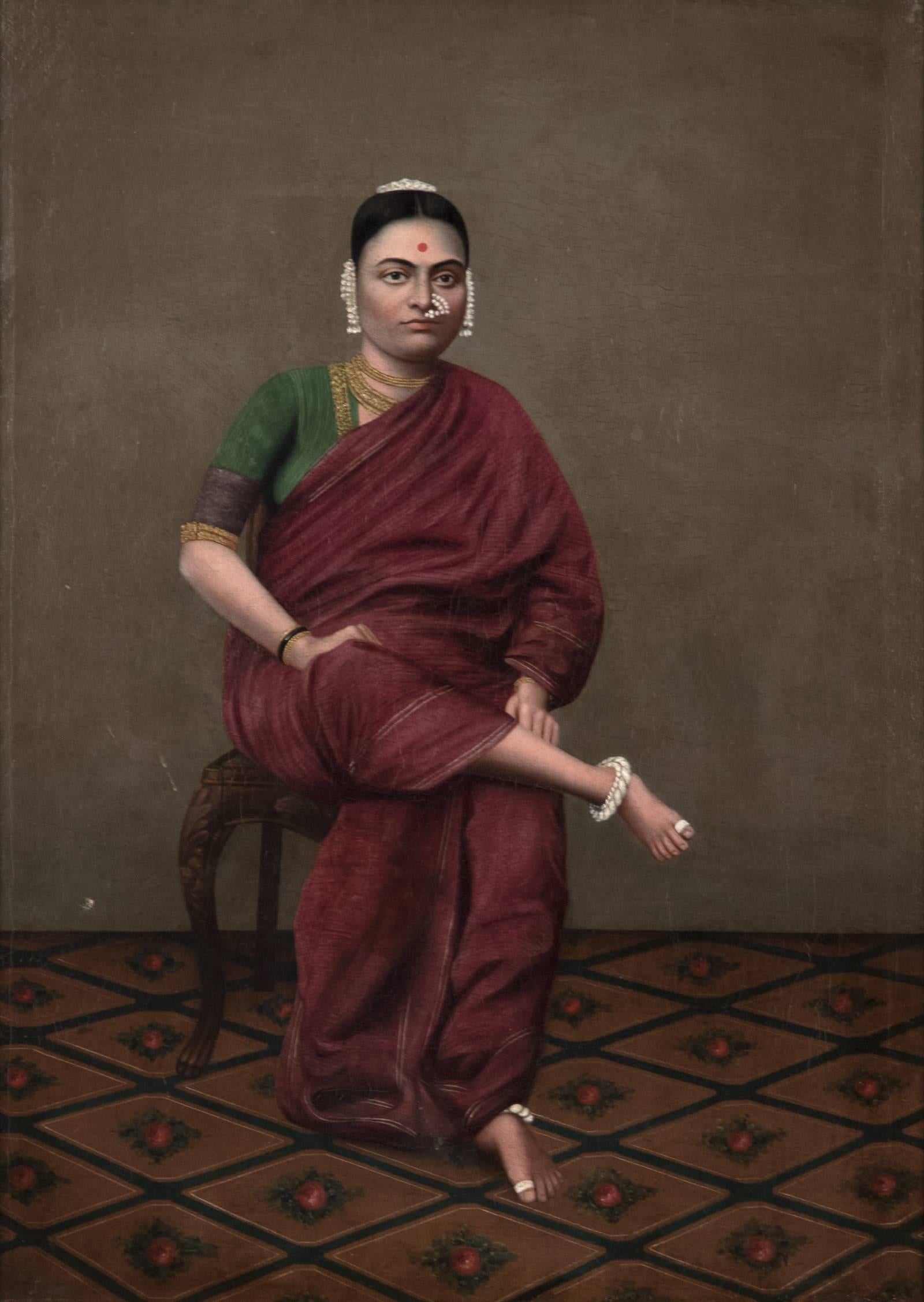 In this 19th century portrait, a woman sits on a carved wooden chair, her right leg crossed over left and her hands in her lap. She is dressed in a gold-trimmed green and maroon sari and is adorned with jewellery; she is a bride on her wedding day.