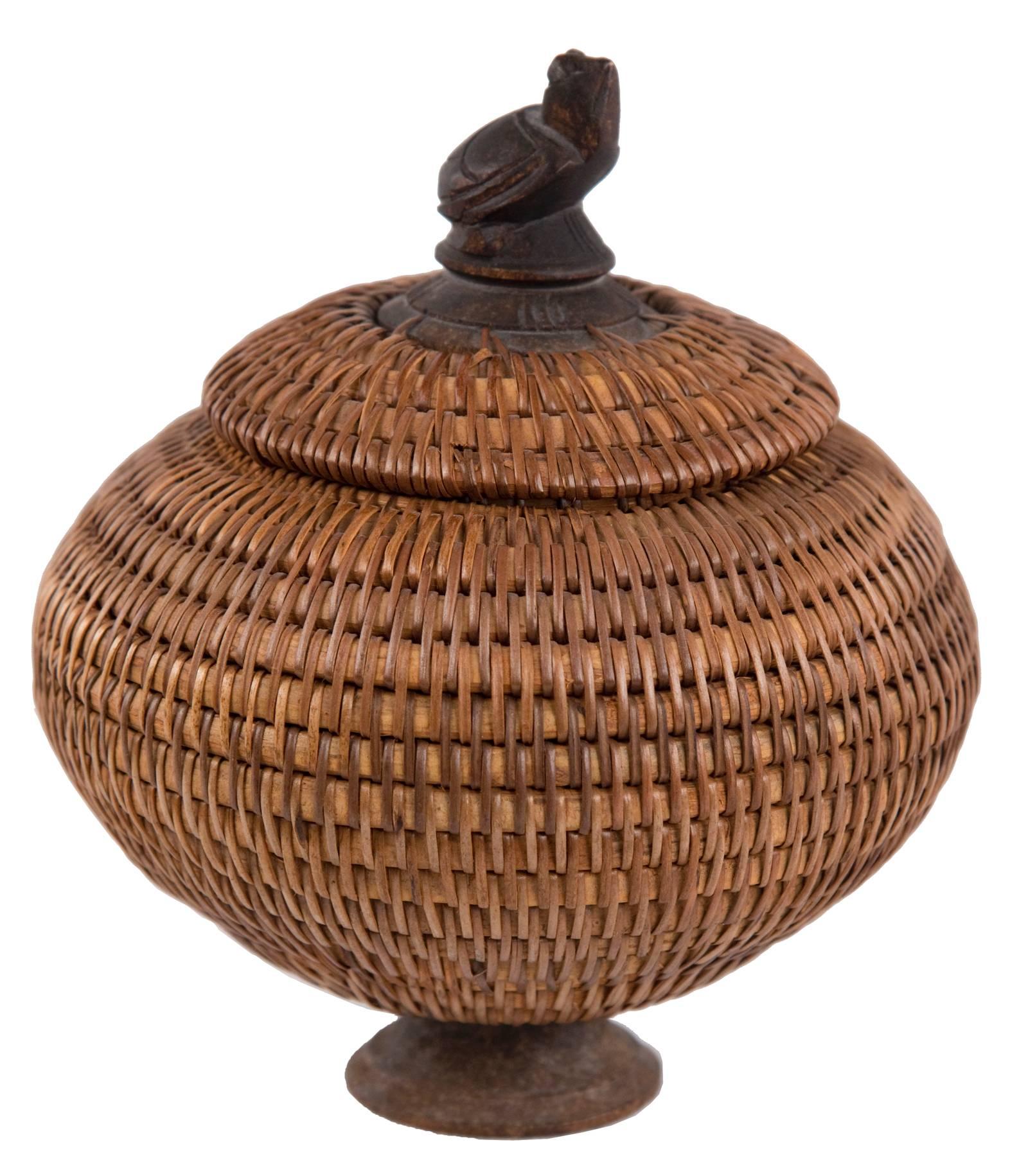 A tightly woven vessel of ovoid form on a wooden circular base and topped with a woven lid with a decorative carved turtle finial.