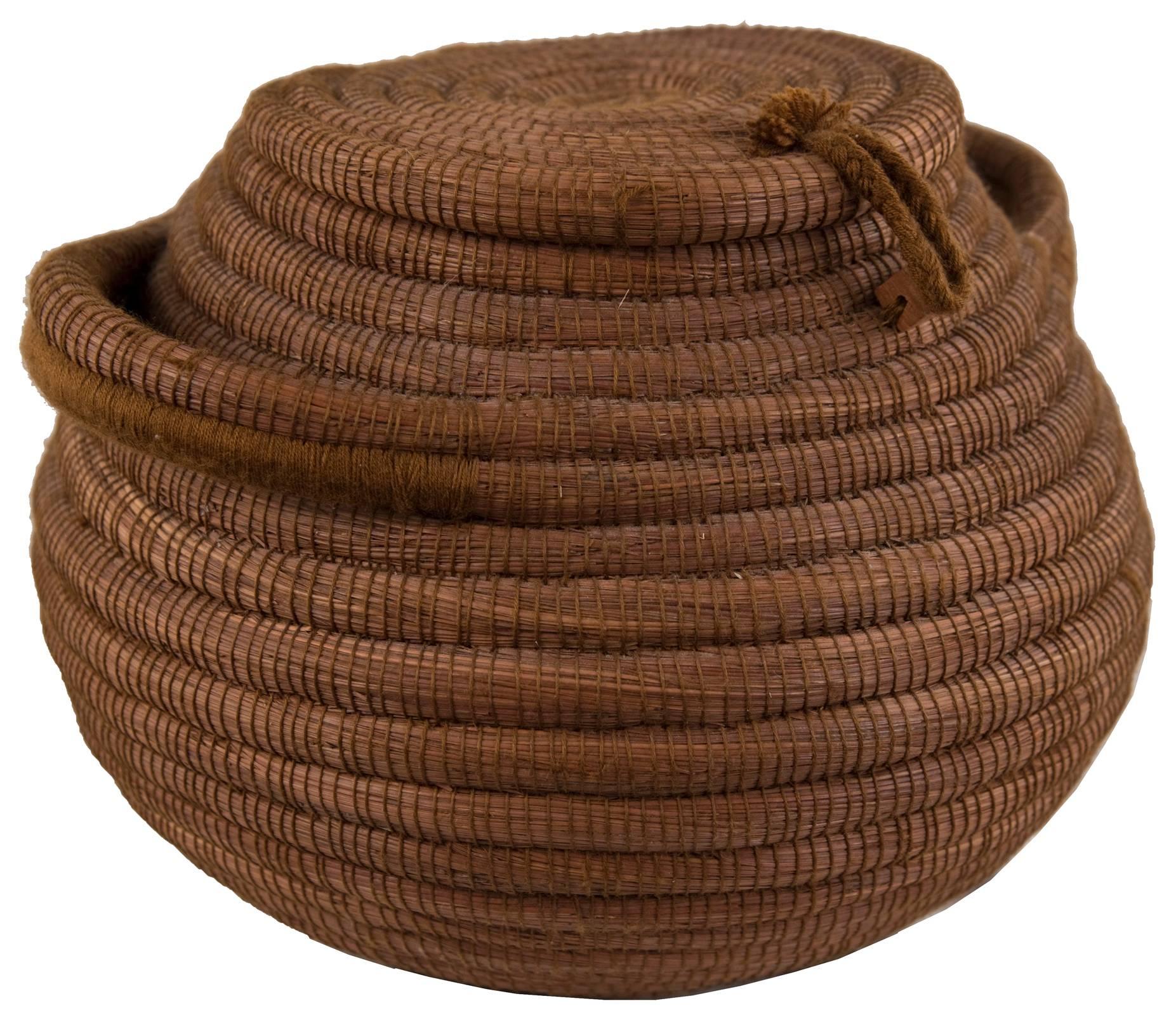Early 19th century papago woven coil basket, woven from pine needles, with hinged lid above a spreading globular-form shape with double body handles, which have been woven into body structure for support. Constructed of woven pine needles.