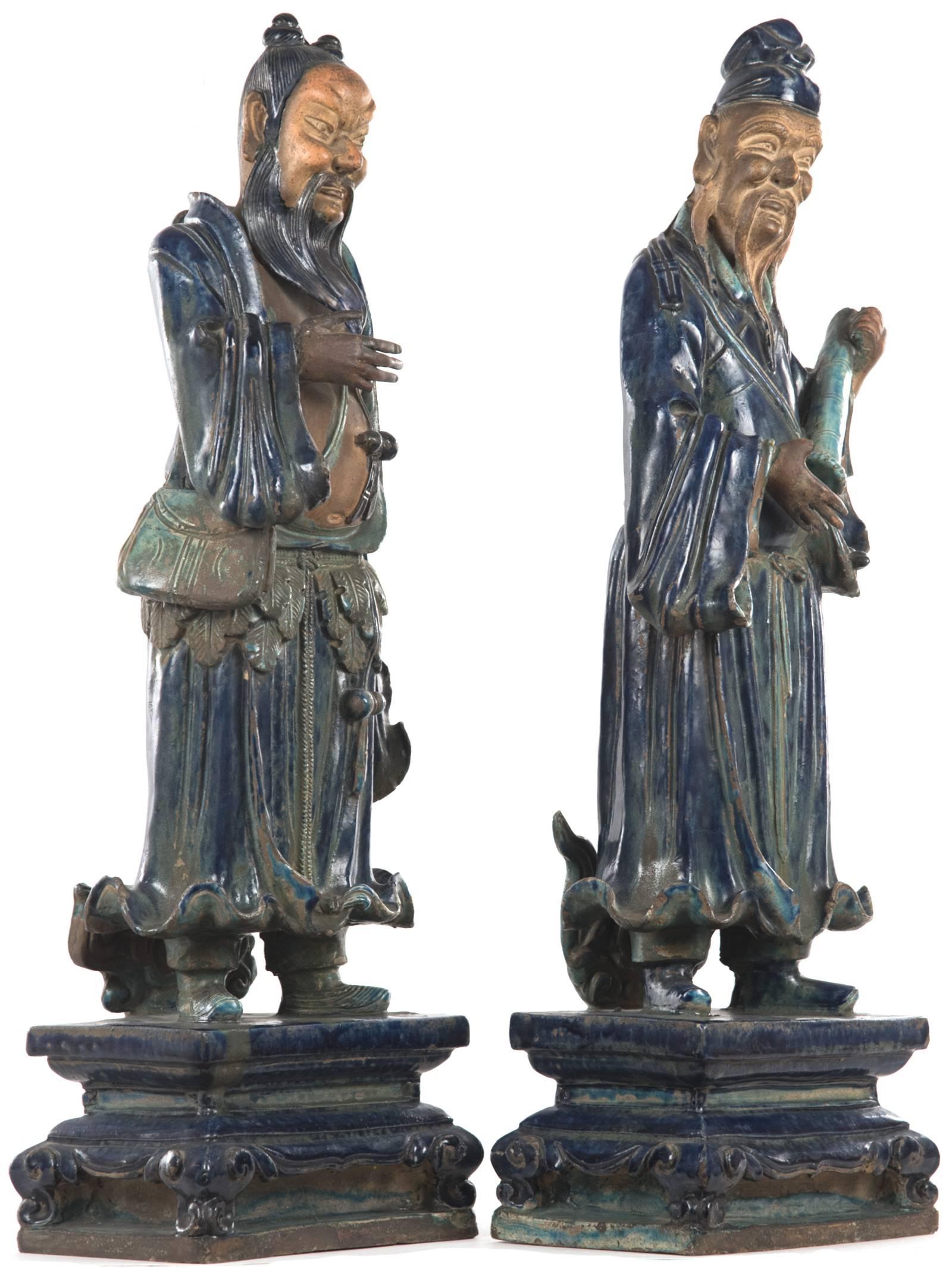 These Japanese immortal god statues are carved from terracotta and glazed in a rich cobalt blue and turquoise enamels, dressed in traditional robes with stylized hems that reveal their shoes, they Stand on short pedestals with a stepped base raised