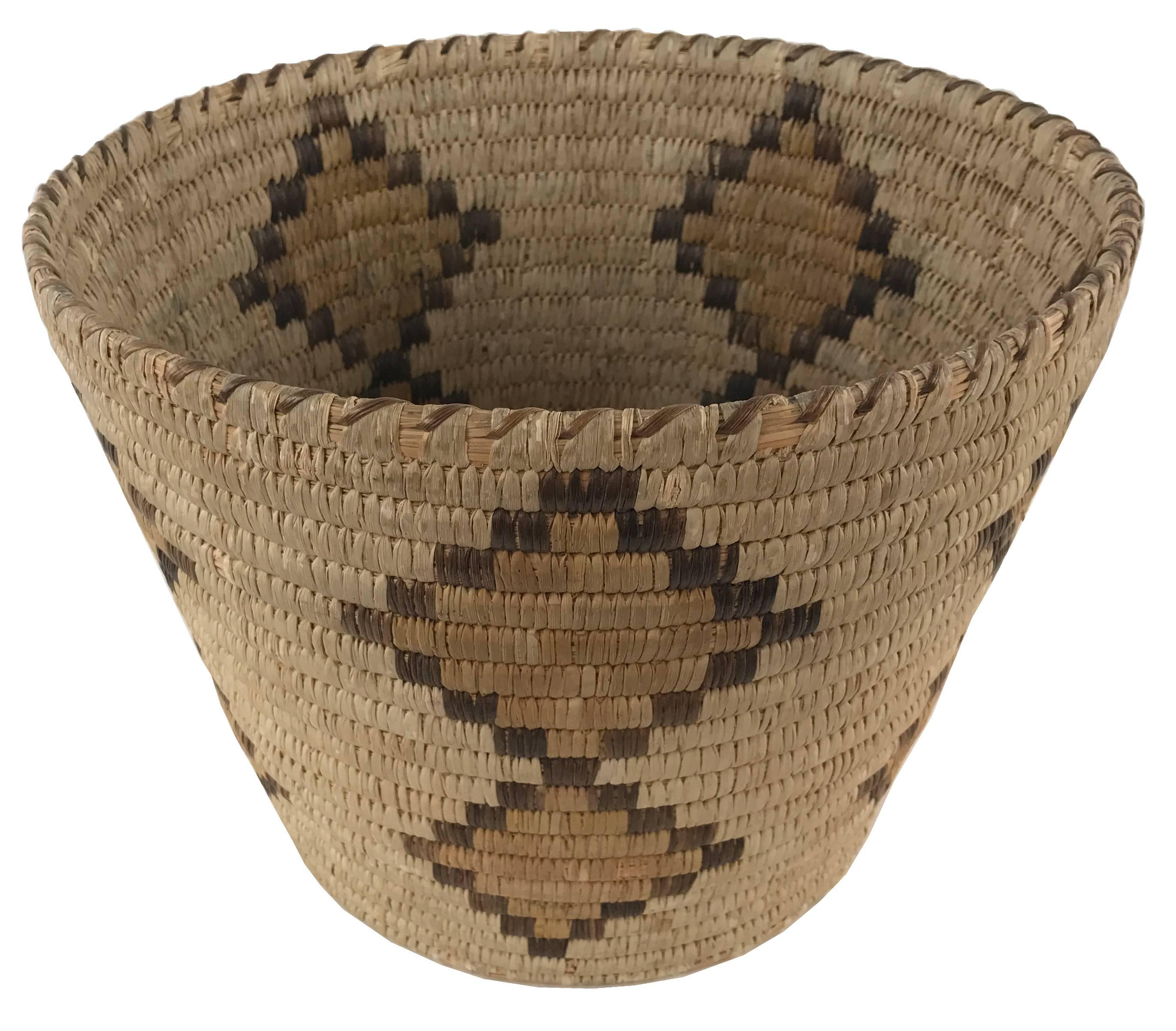 A Native American twined woven basket with a tapering form leading to a coiled base. The body is decorated with a woven double diamond motif in a dark and yellow design that contrasts with the light ground.