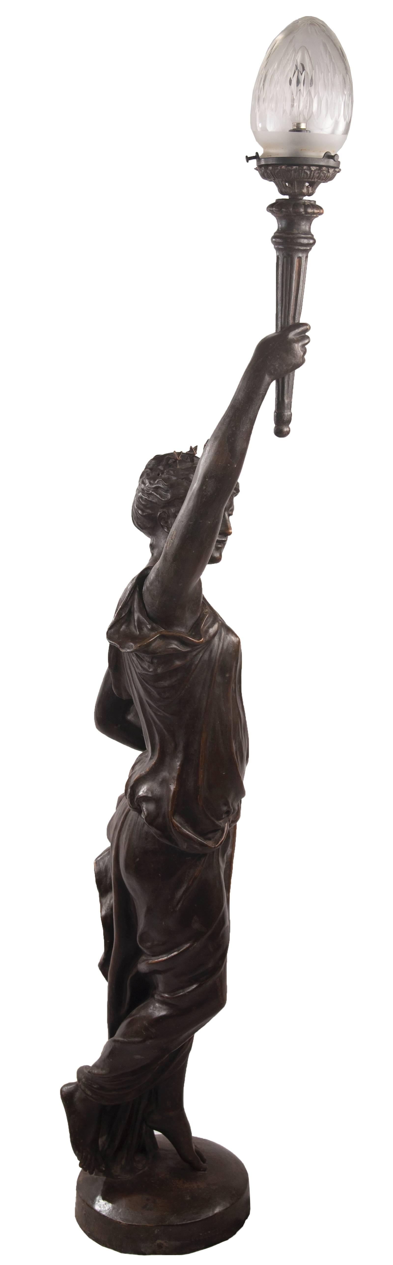 A figurative patinated bronze floor lamp perhaps depicting a representation of Lady Liberty supported on a round base. A six-star crown rests on her head while one hand draws back her robes as to not interfere with her sense of determined motion,