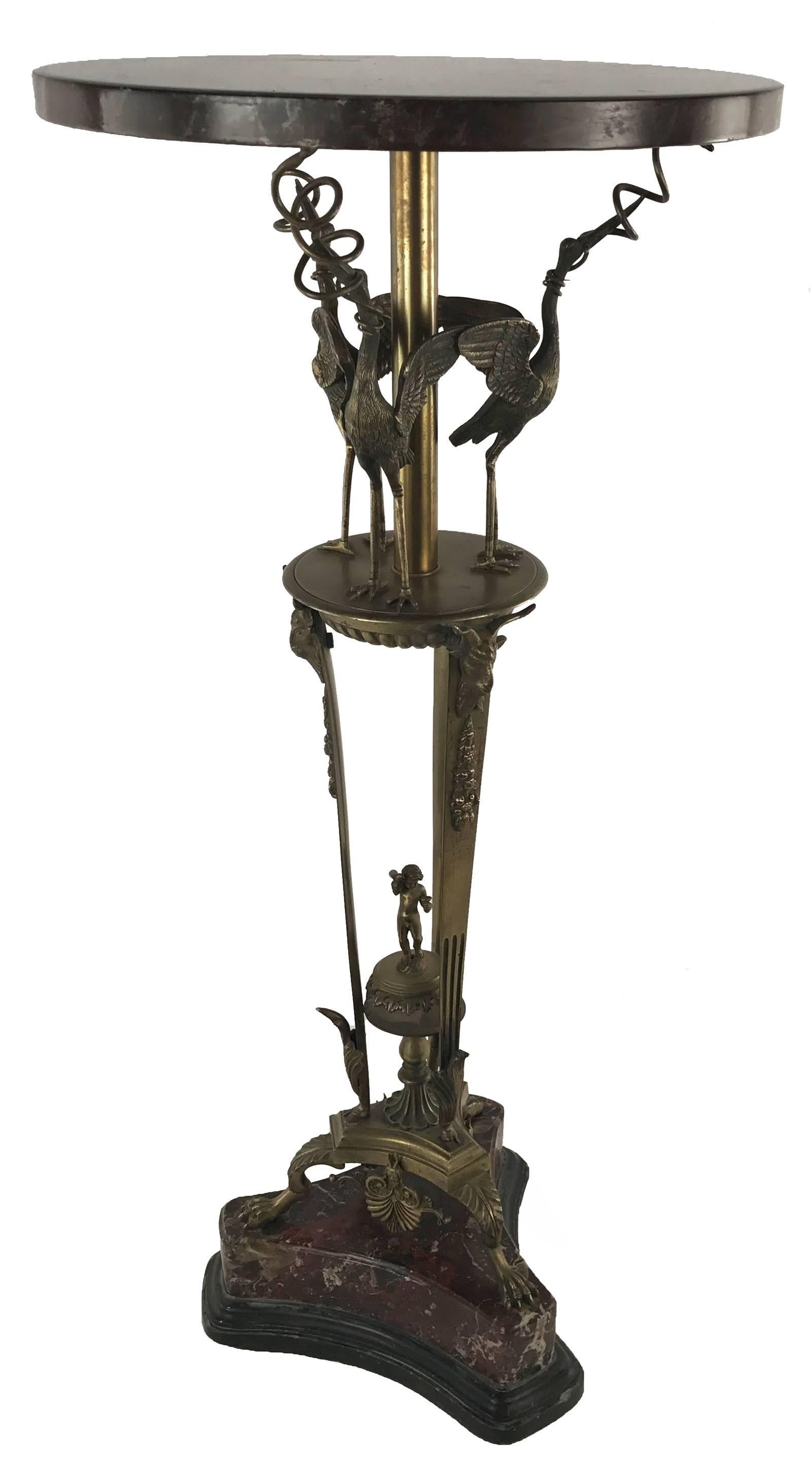 An Empire style pedestal table with an acrylic, red faux-marble top that surmounts a bronze stem and round base on which are three standing cranes with spirals that support the tabletop. Three bronze legs with applied bacchus and garland motifs are