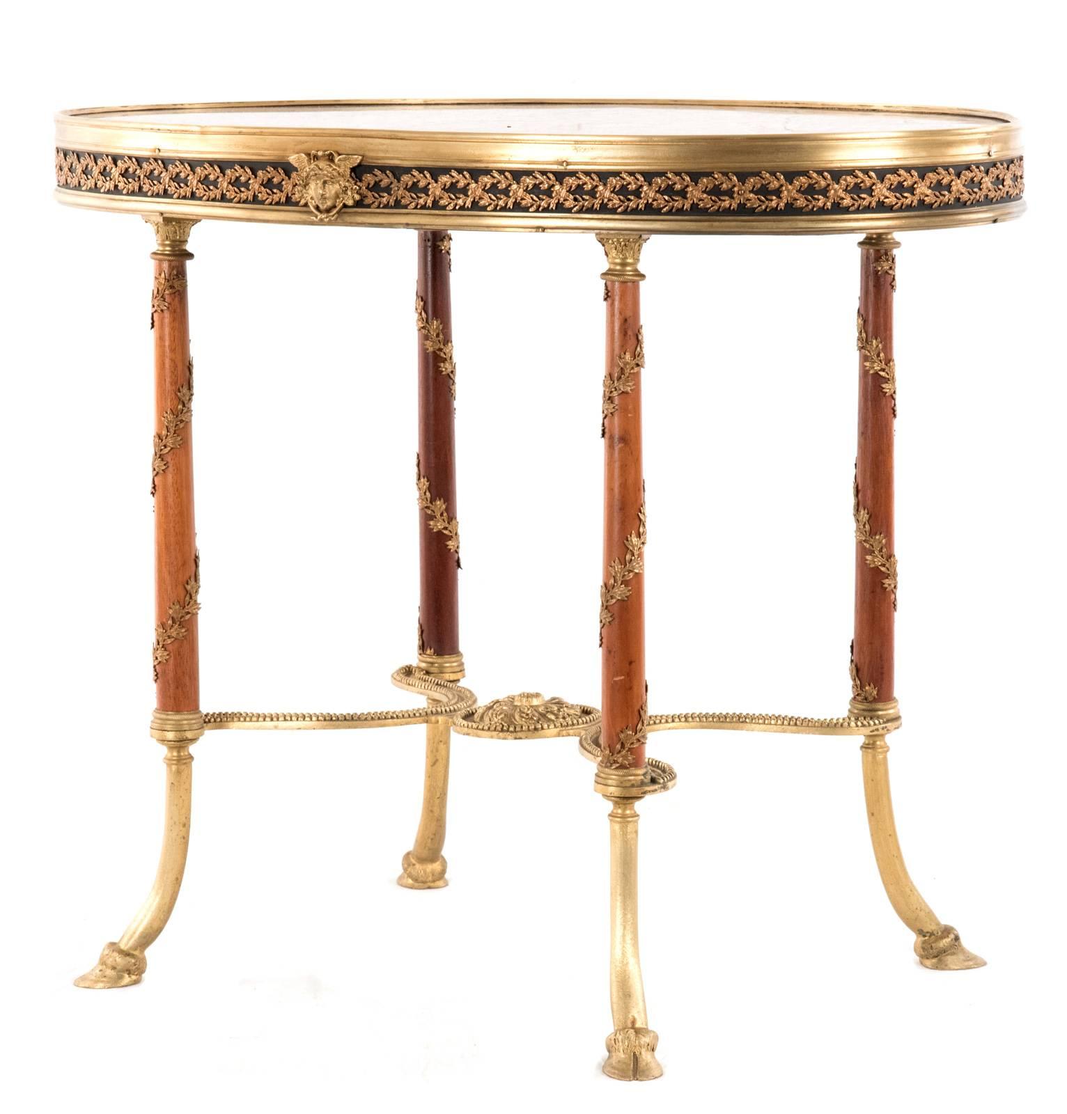 French oval marble top sitting on a finely cast brass gallery border with vine detailing supported by four column-shaped legs wrapped with leaf and vine carvings raised on slightly splayed hoof feet, which are connected by an X-frame stretcher with