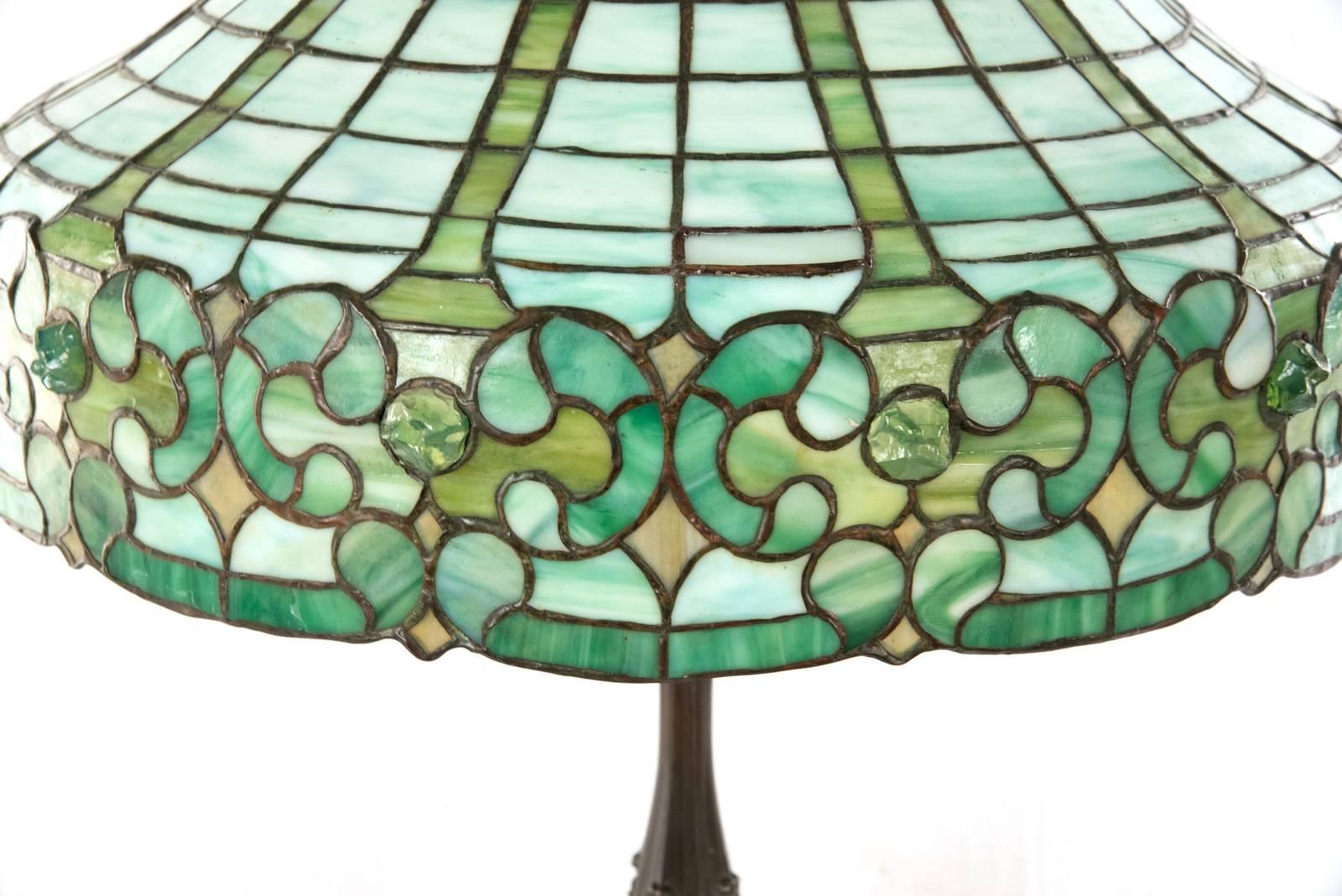Made by the JA Whaley of New York, this handsome leaded glass and bronze table lamp with a faceted shade comprised of individually camed panels of various types of green glass over an apron of corresponding c-scroll and medallion panels, sitting