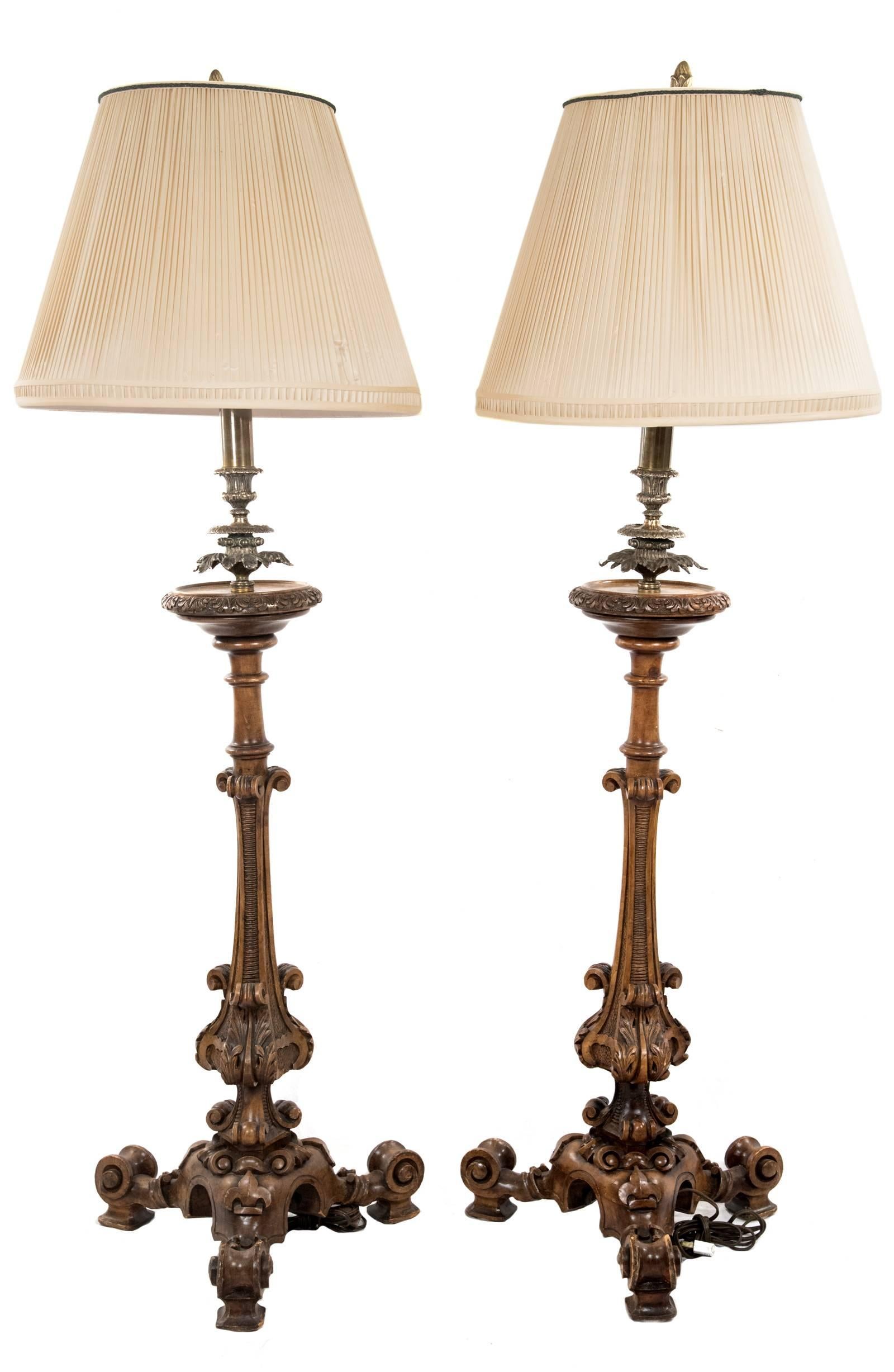 A handsome pair of table lamps with incised and shaped brass neck and collar supported on a carved wood base with scroll work and stippled decoration above unfurling acanthus leaves, all raised on an ornately carved pedestal base with square feet.