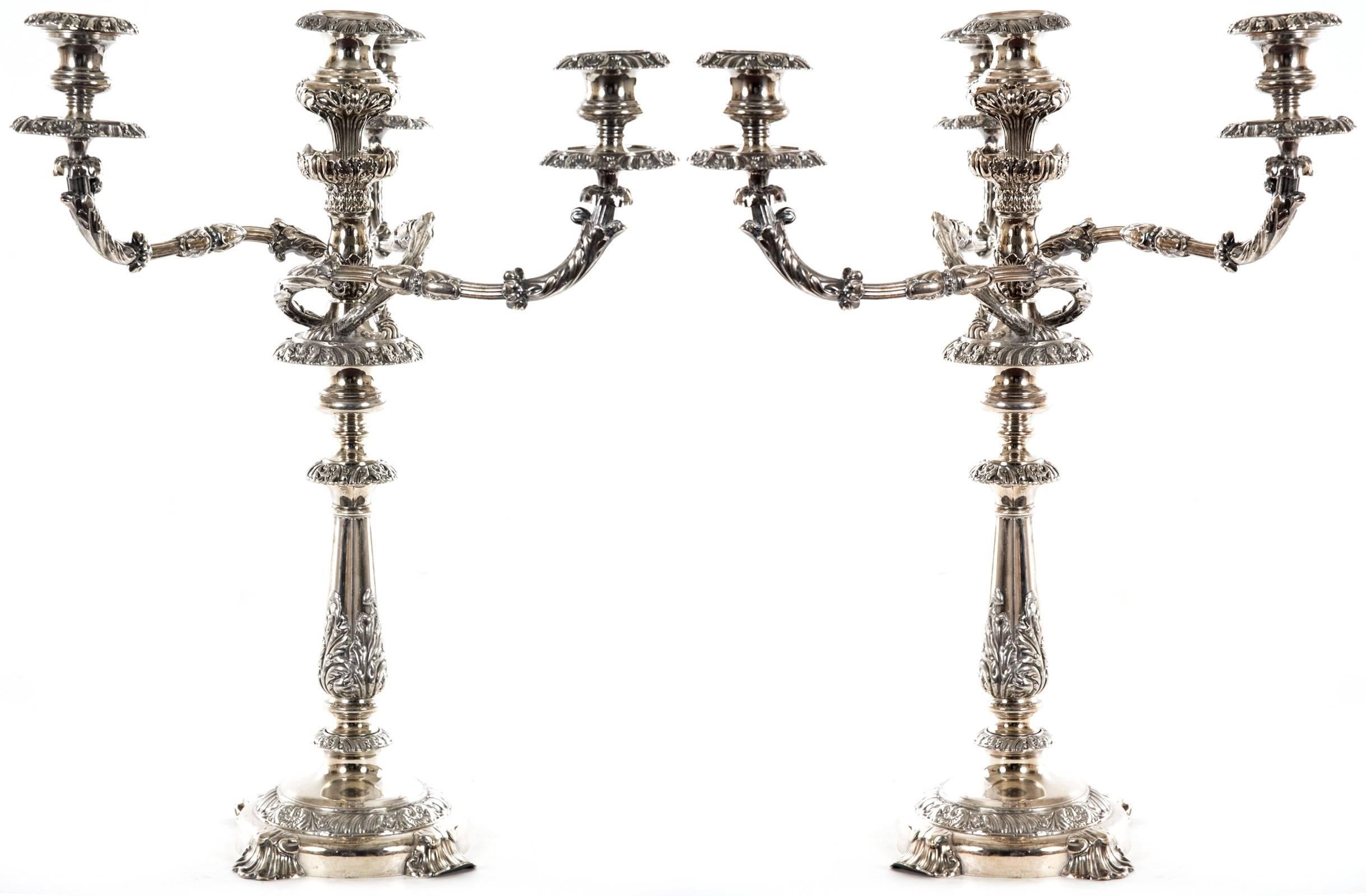 A pair of silver plated candelabra with three branches, as well as a sconce atop the centre stem, embossed with scrolling leaf and foliate decoration, supported on a round base with four applied foliate feet.
