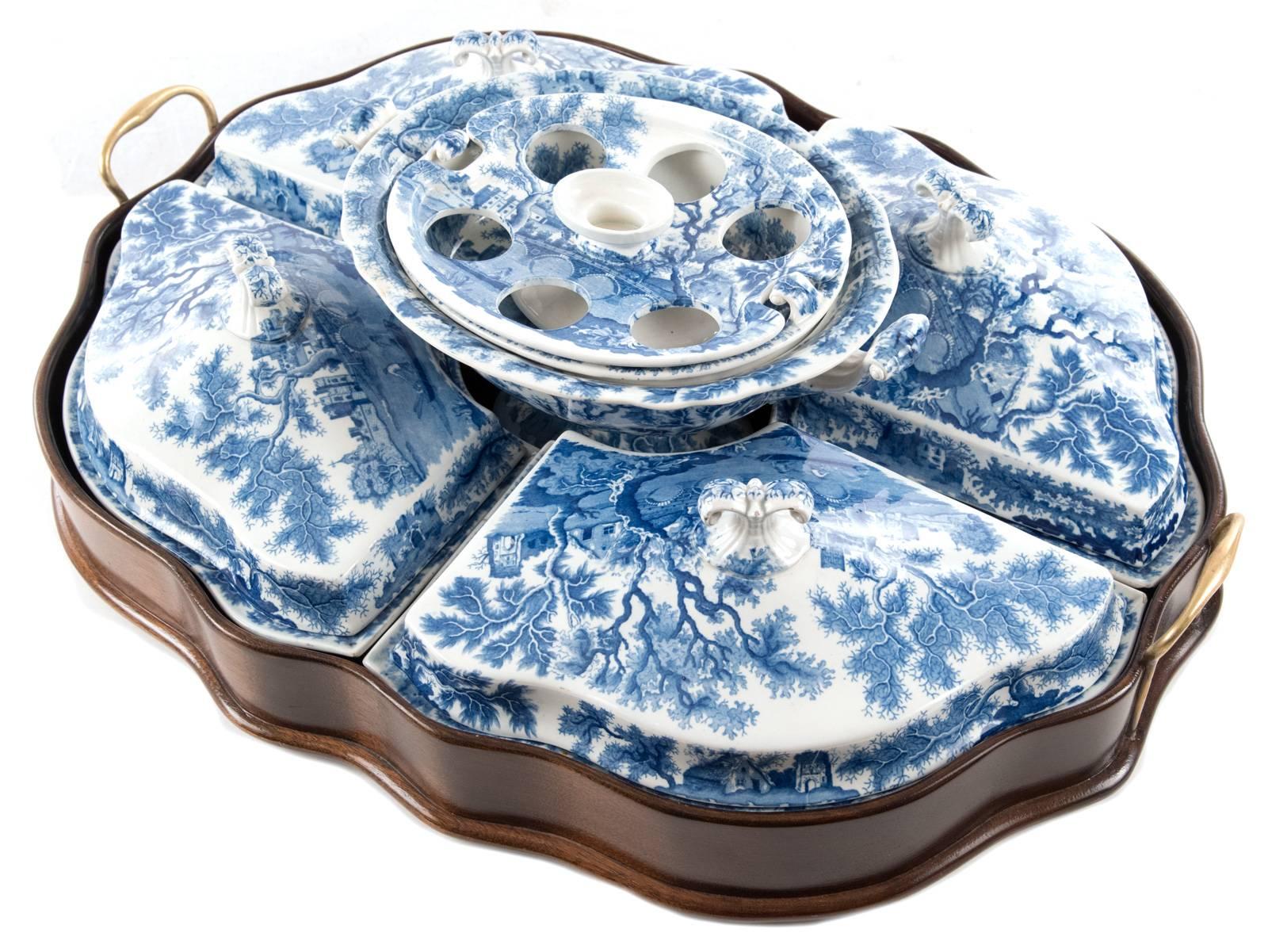 A double-handled brass serving dish of scallop form inset with blue and white porcelain serving compartments, which comprise four large dishes with lids around a central tureen, all decorated with a transfer design of an idyllic view across a river