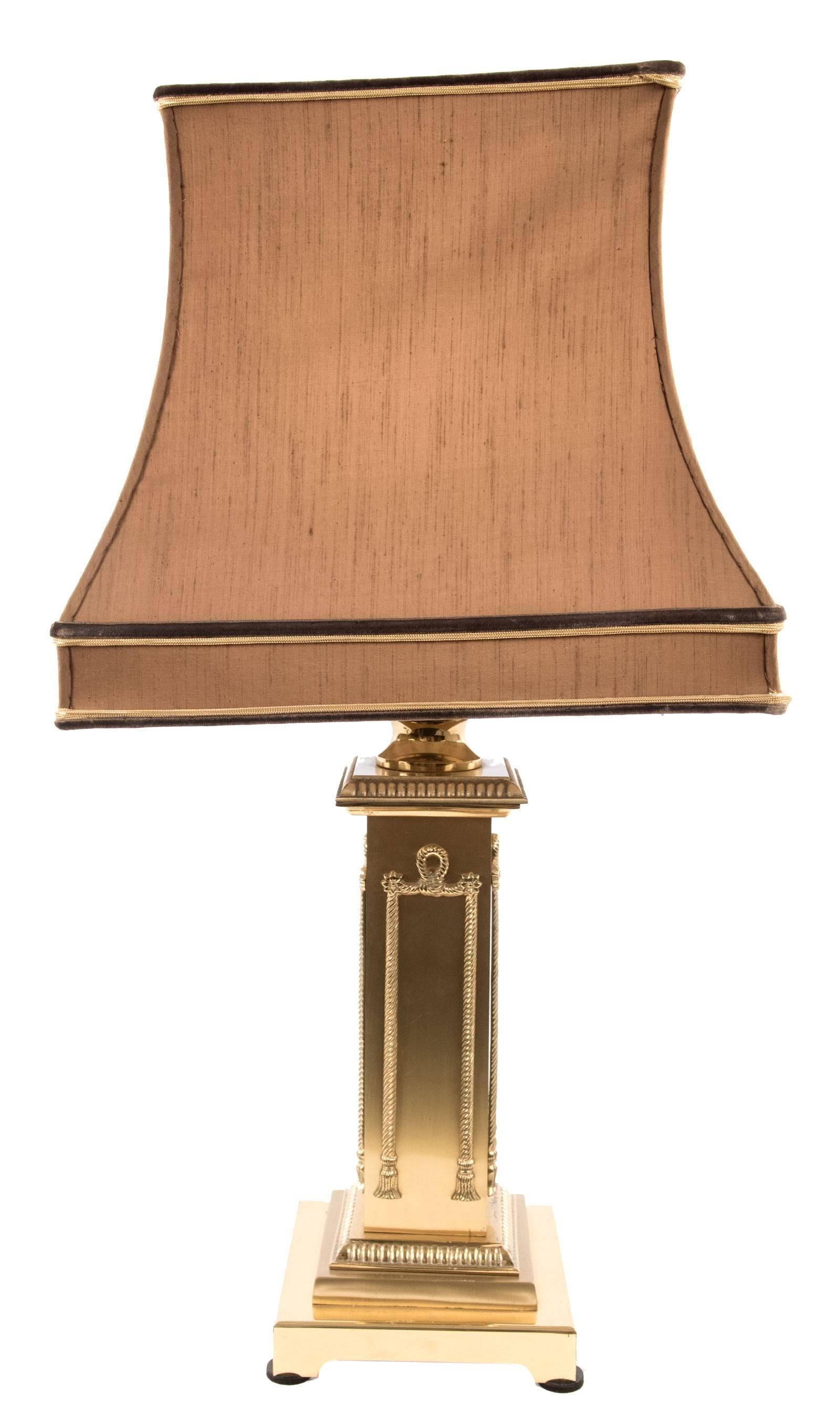 A gold-plated table lamp with an elongated rectangular form that is decorated on all sides with a stylized rope hung by floral medallions and is flanked above and below with raised gadroon trim. The lamp is raised on a square stepped base, and the