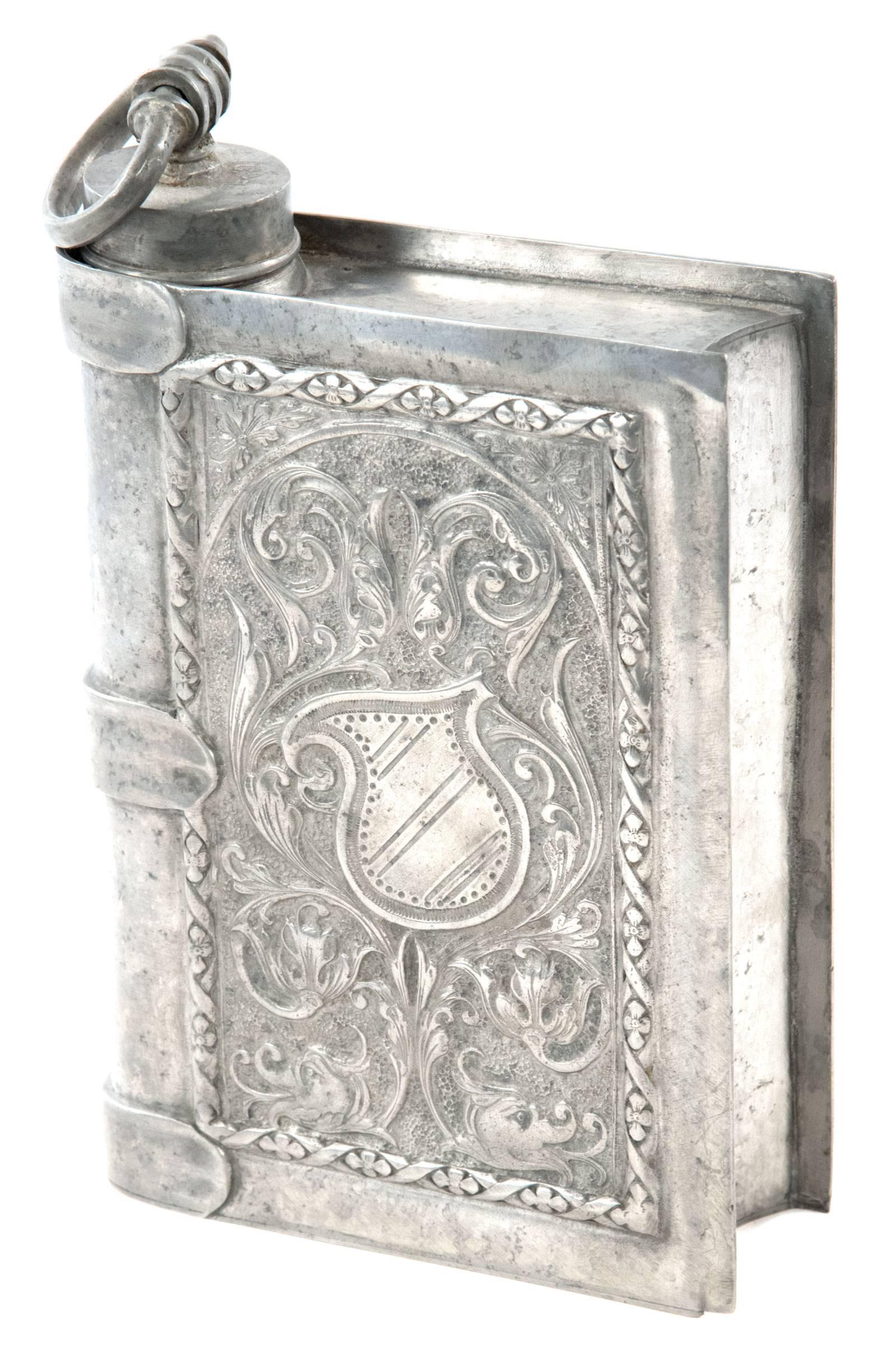 A pewter flask taking the form of a leather bound book with a banded spine and elaborately decorated covers. The front with a design of scrolling florals and foliate around an escutcheon centrepiece, all of which is surrounded by a raised border of