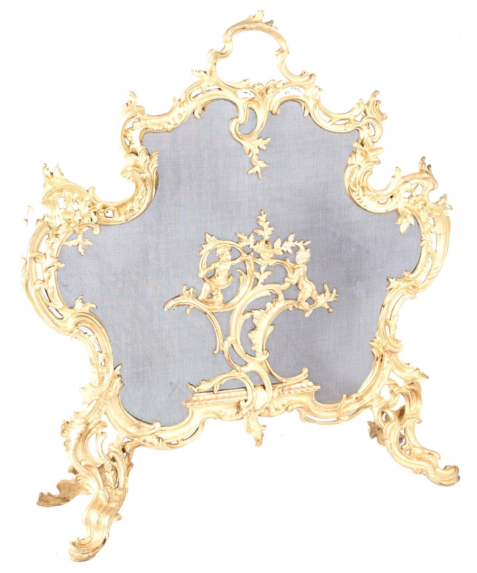 A French gilt-brass Rococo-style fireplace screen, the ornate frame with scrolling foliage and filigree designs and a pair of cherubs on the screen, raised on scrolled and pierced legs.