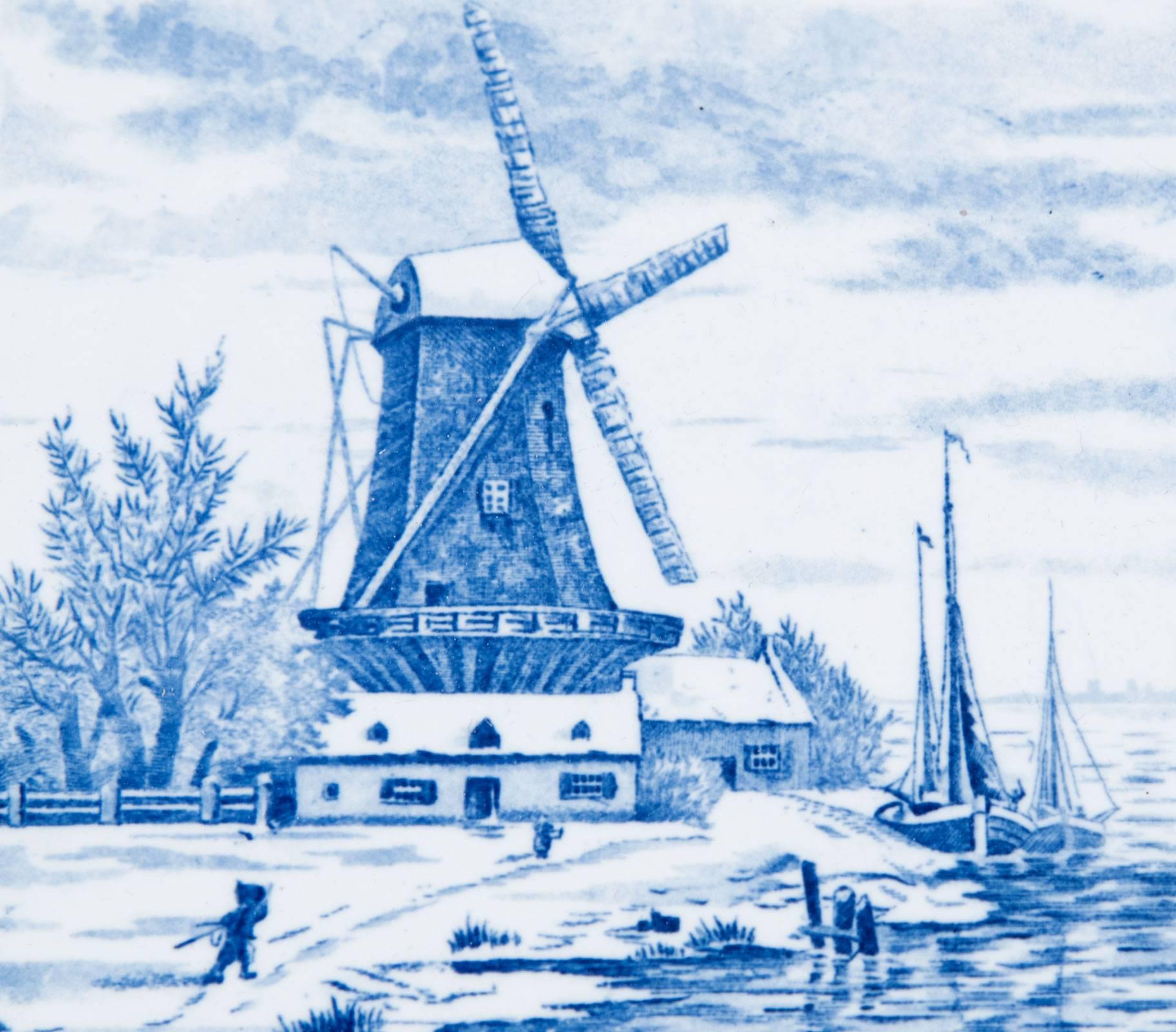 A Delft style blue and white tile depicting two figures walking along the shore of a waterway upon which sits a large windmill among architectural buildings and trees. Hand-painted with rich cobalt blue pigment, this large tin-glazed-earthenware