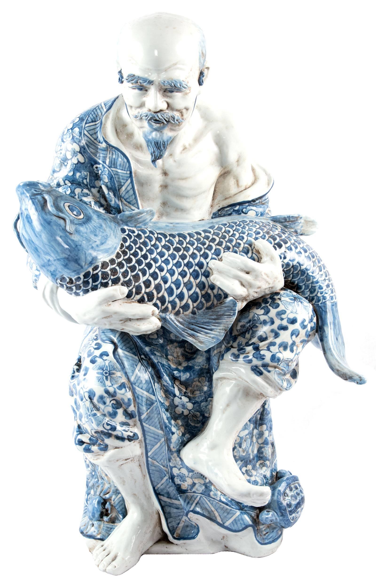 A blue and white painted porcelain figure of a man struggling to carry a large koi fish in his arms, his beautifully decorated robe falling from his shoulder and his facial expression shaped with the effort. Dimensions: 27 x 16.5 x 9.5 in.