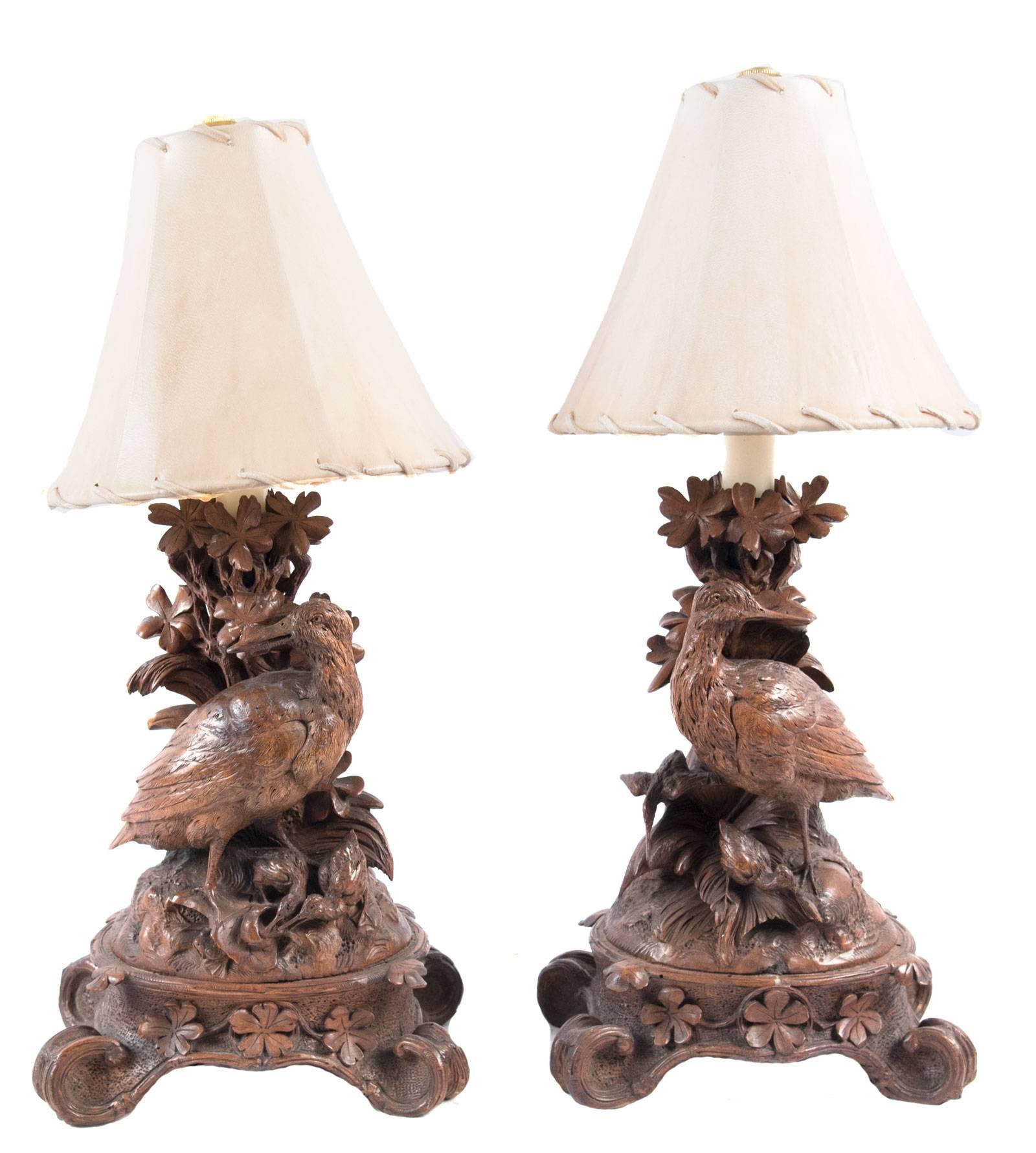 A pair of elaborately carved wood table lamps in the Black Forest style popular in the late nineteenth century. Opposing pheasants are perched on rocky ground, against a trunk, foliage and pinecones, the figures on a similarly carved base and topped