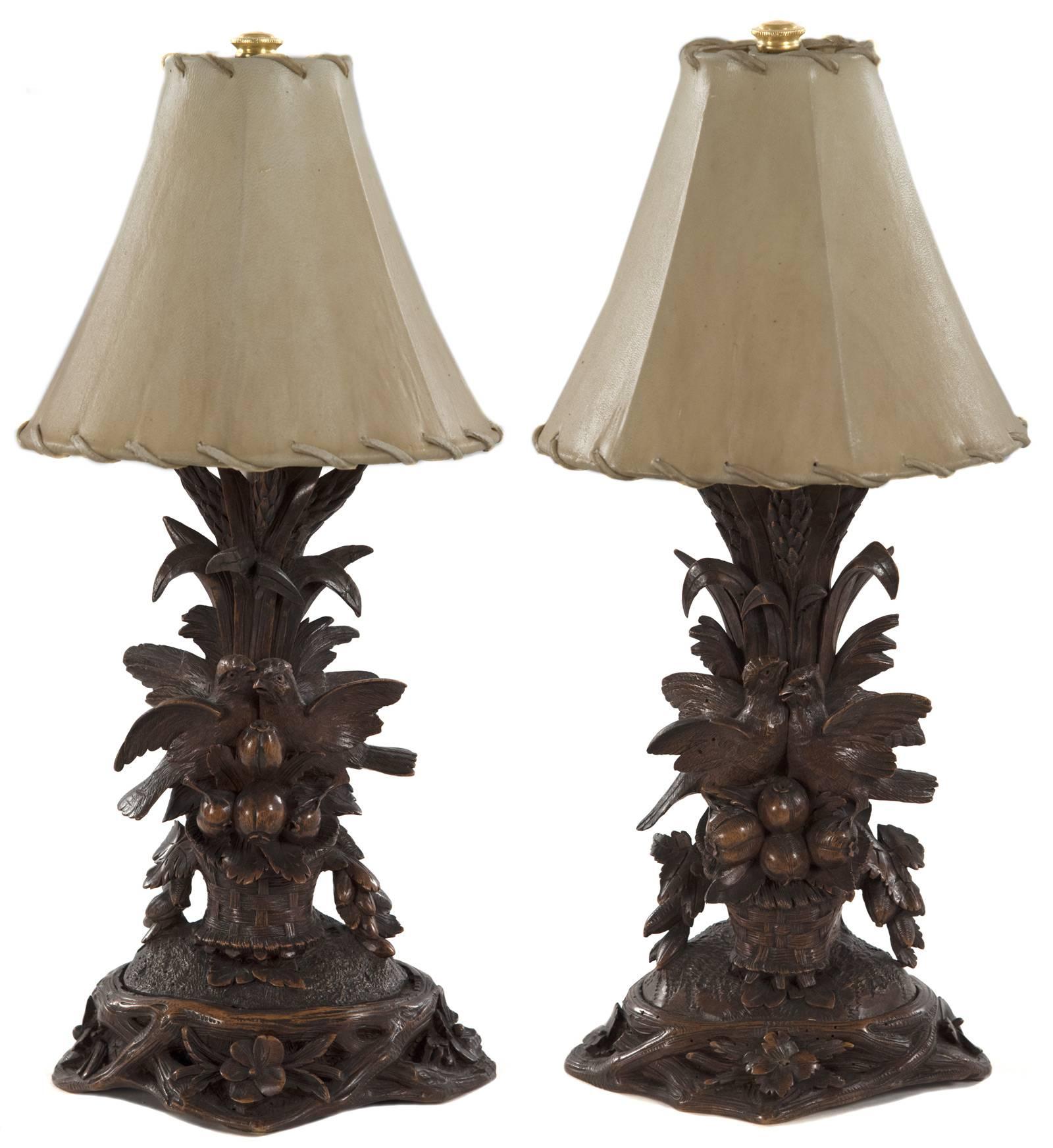 A pair of elaborately carved wood table lamps in the Black Forest style popular in the late 19th century. In front of tall stalks of grasses, pairs of birds are perched upon a small bushel of persimmons and foliage that sits within a woven basket on