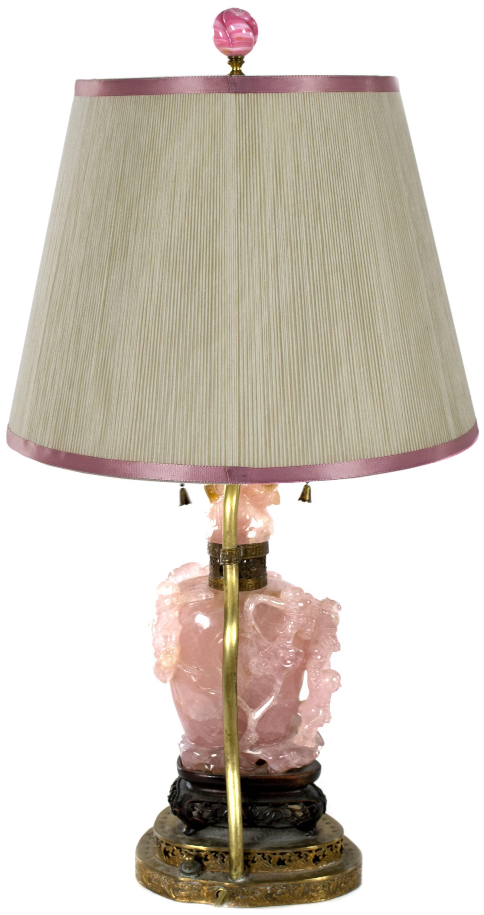 Set on an ormolu stand, this rose quartz table lamp is carved with various figures, shells, and foliage. In addition to the two light bulbs beneath the shade, the interior is lit with another light.