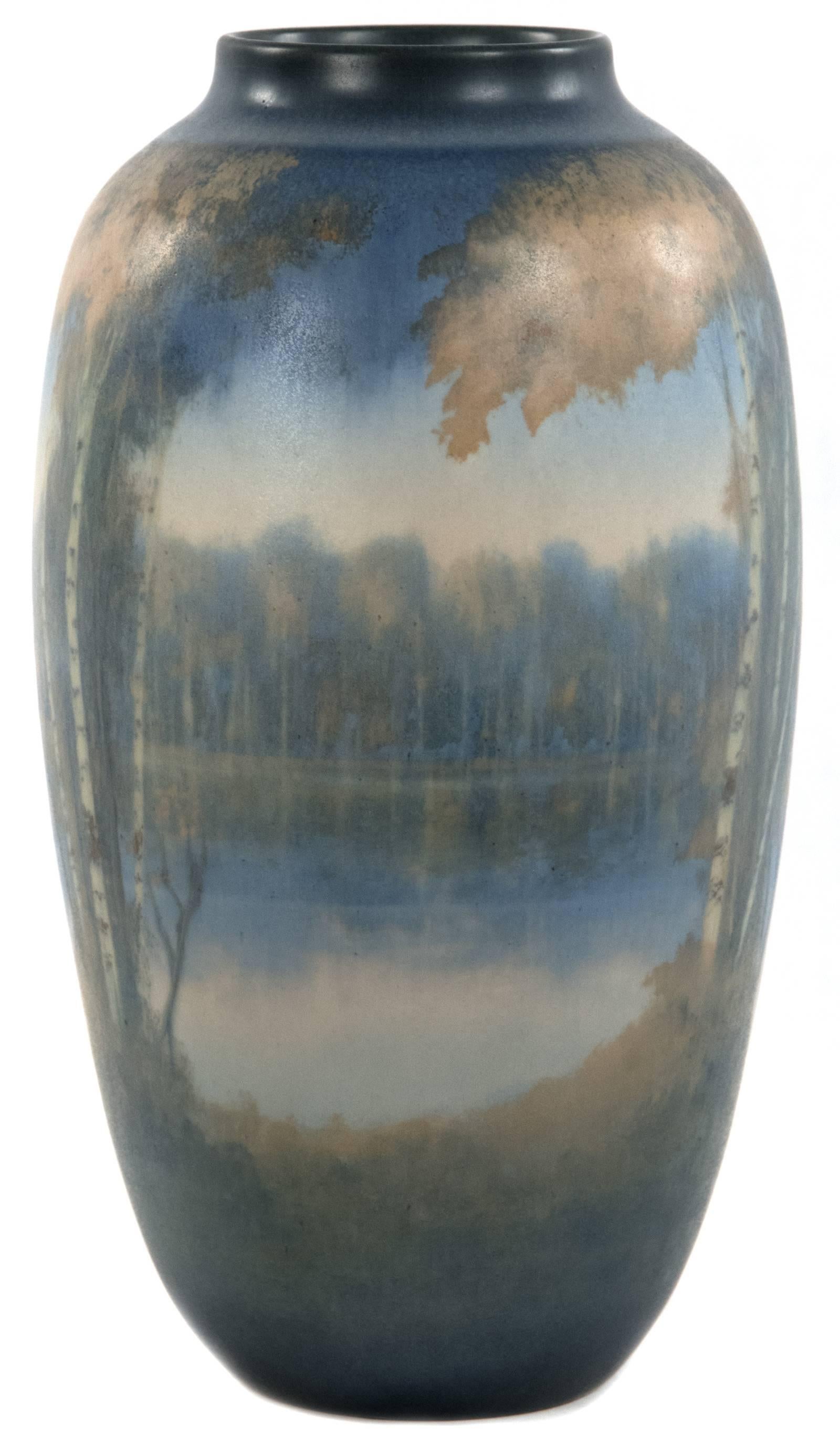 A tree-lined shoreline is reflected in a calm river in this atmospheric landscape that is softly illustrated in a neutral autumnal palette of blue, orange and brown glazes on this Rookwood Pottery vase of ovoid form dating from the mid-20th century.