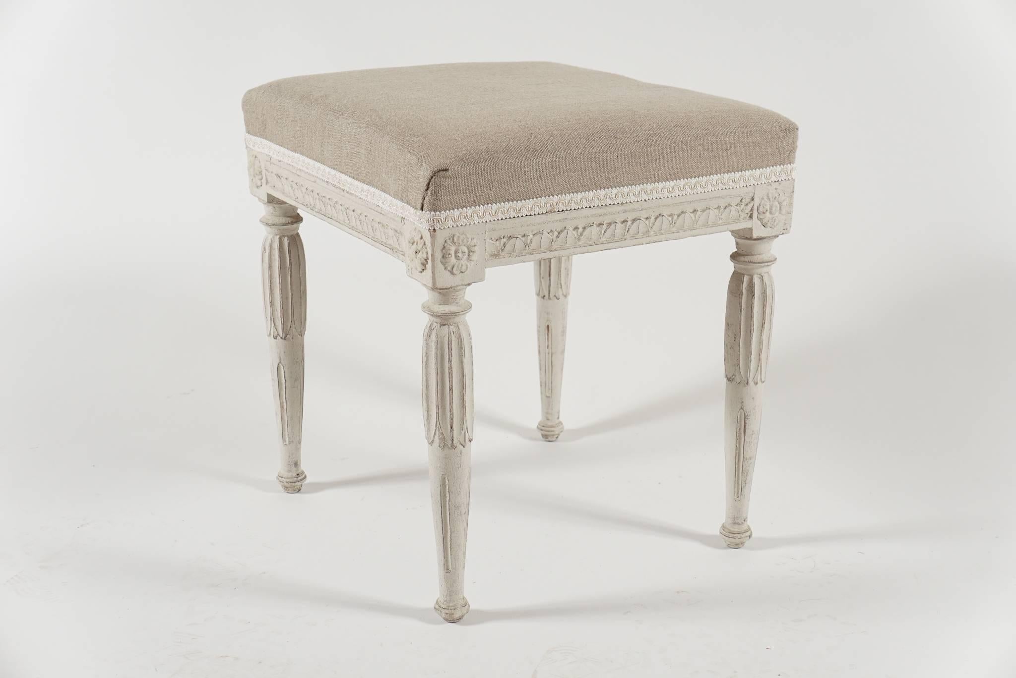 A near pair of circa 1790, Swedish Gustavian period wood stools in original paint having upholstered seats above carved 'lamb's tongue' seat rails joining rosette carved corner blocks on fluted, turned, and carved acanthus legs.  One measures 16.5