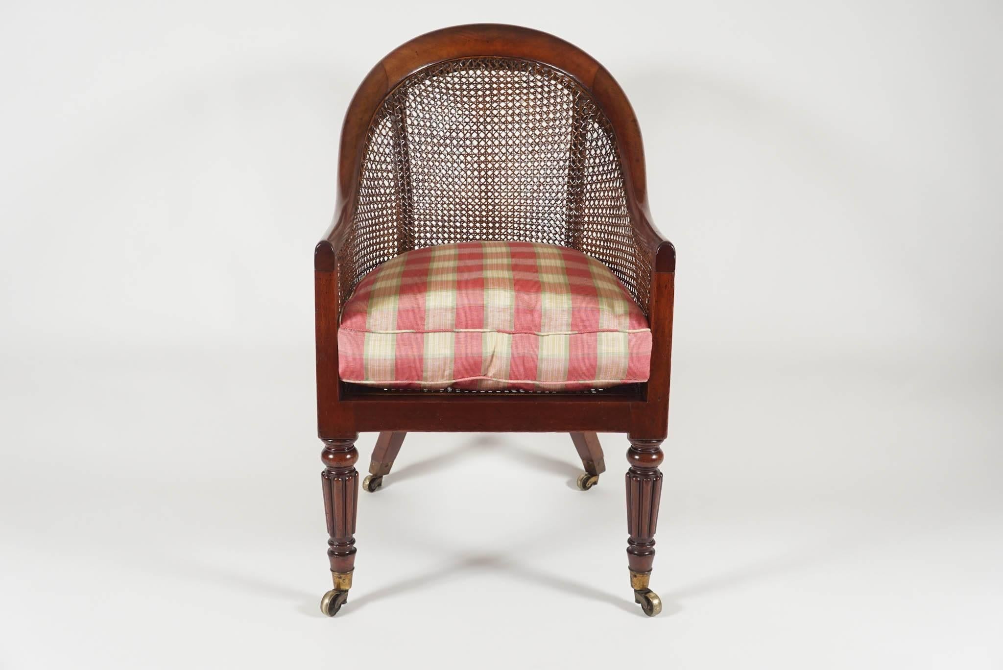 An elegant English Regency period, circa 1820, armchair, library chair, or bergere of solid mahogany construction having rounded crest rail with caned back, sides, and seat with loose squab cushion on turned and reeded front legs and splayed rear,