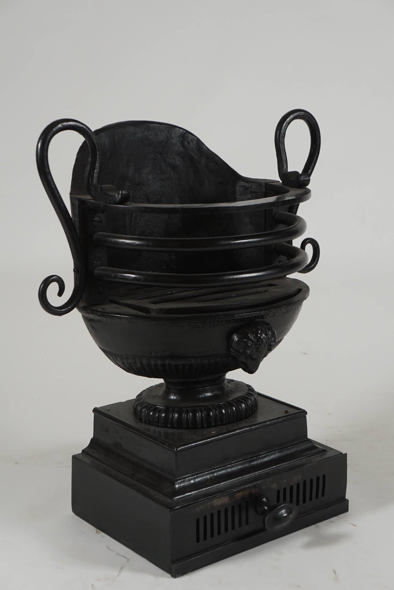 An elegant 19th-century English Regency style cast and wrought iron fireplace coal grate or basket of urn or trophy form having wrought handles either side of cast body with ram's head ornament on plinth with integral ash drawer and oval knob