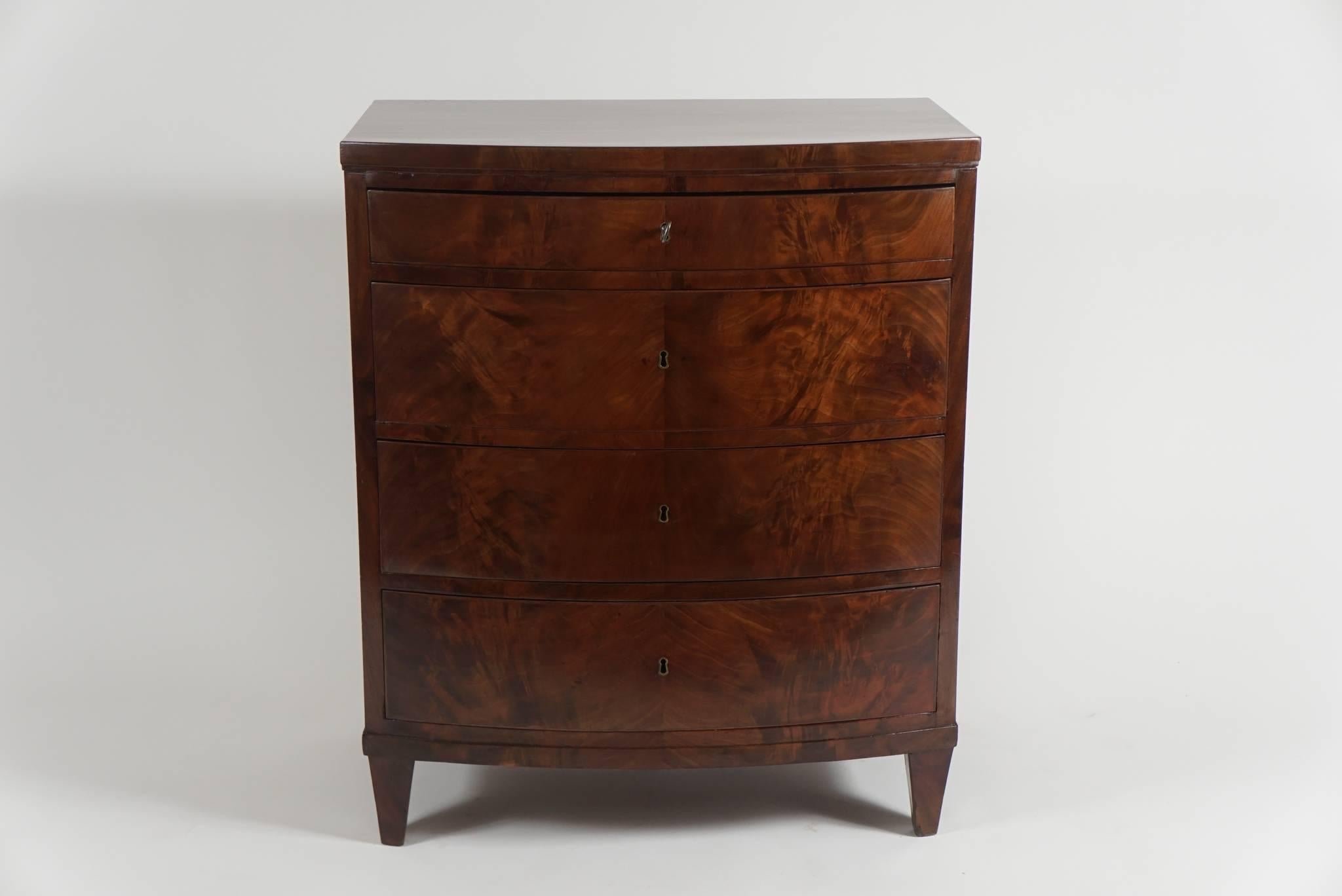 An elegant, richly grained mahogany bachelor's chest or petite commode, of bow front form and clean design having four drawers with central locks on four tapered supports. Considered 'Empire' due date of construction and the luxurious use of