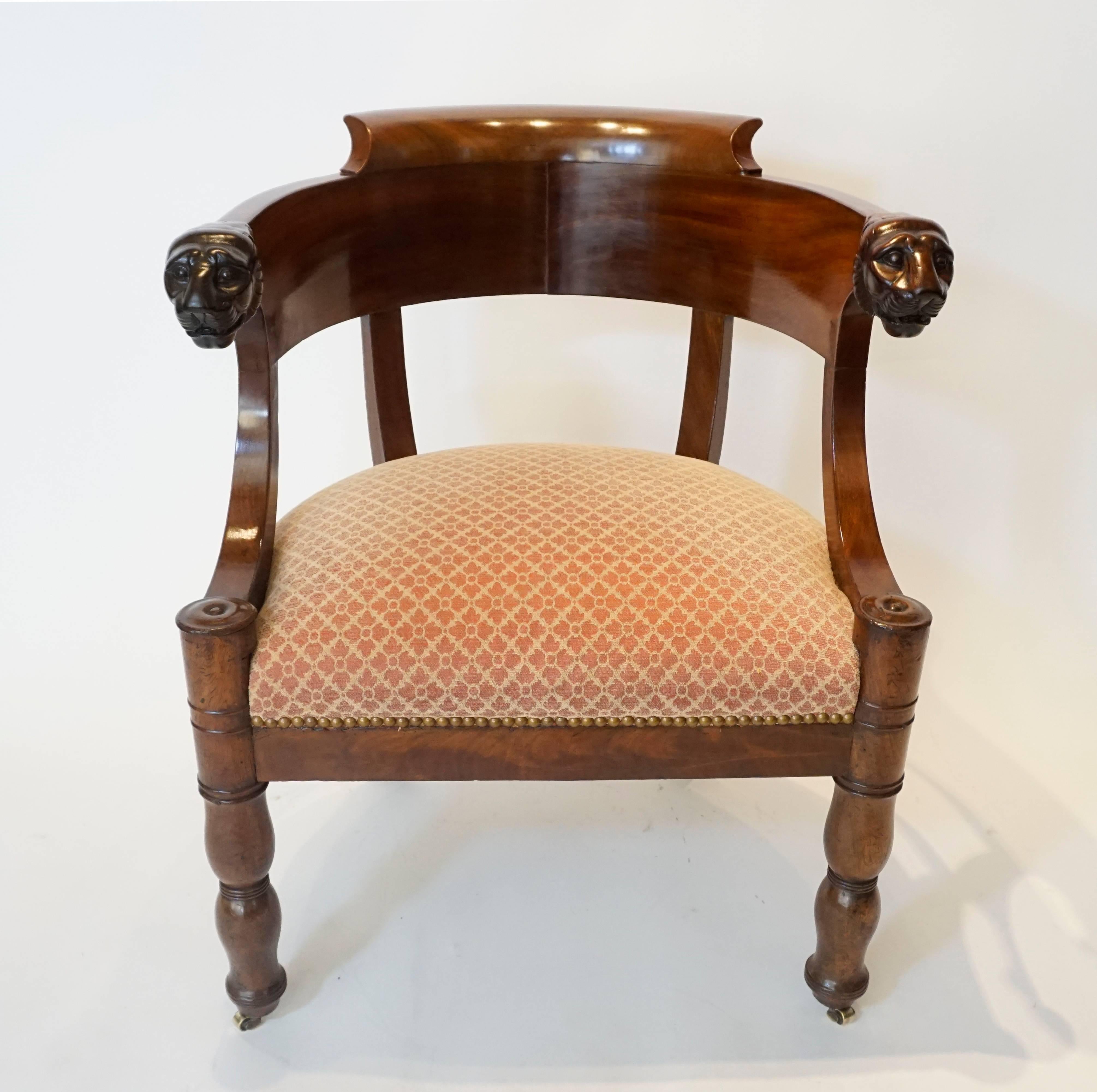 An elegant French Empire, circa 1810, fauteuil de bureau, or desk chair, having carved mahogany frame with continous deeply curved klismos-influenced backrest terminating into finely carved lion-muzzles; swept arm supports joining round turned front