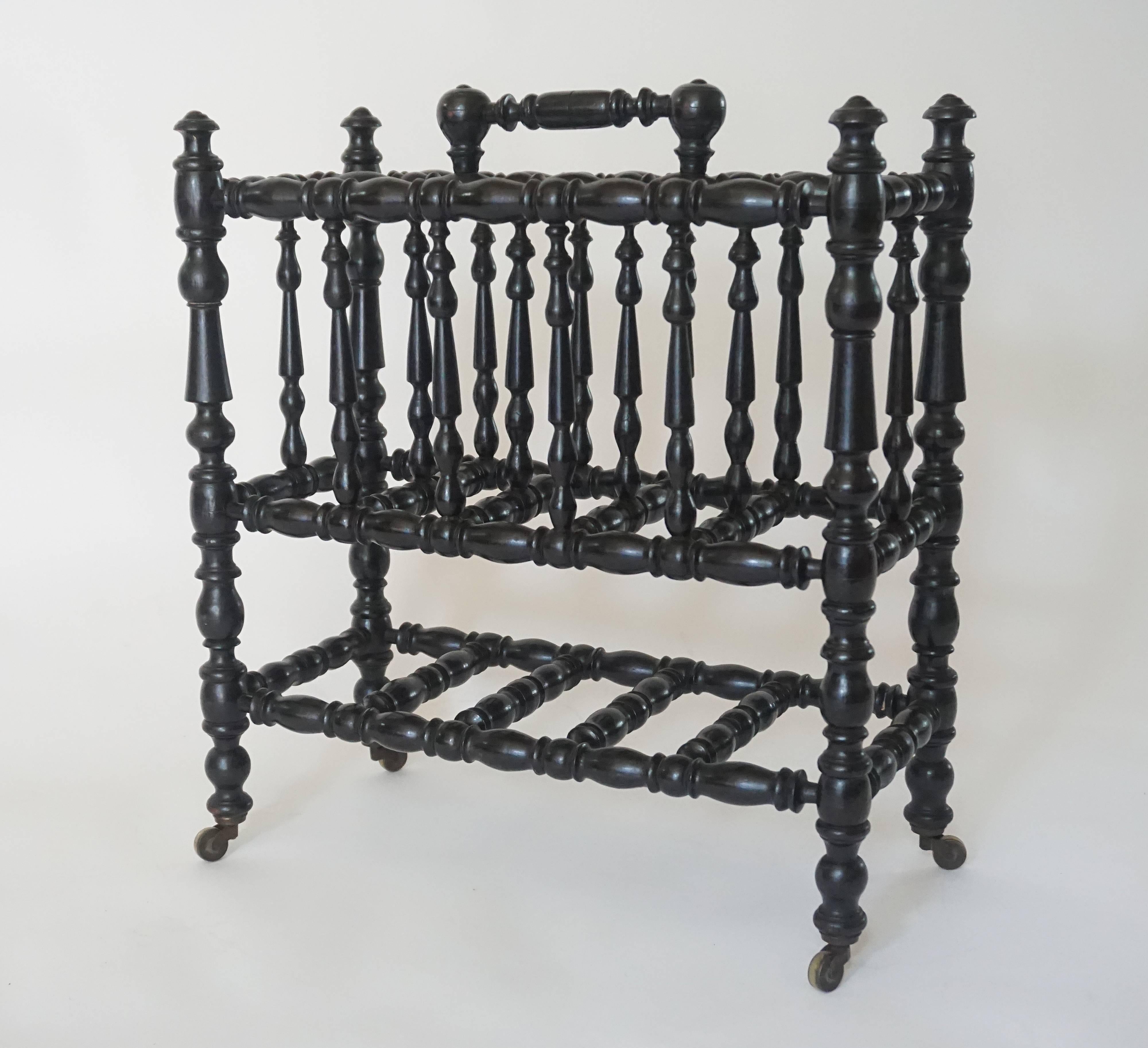 Anglo-Indian portable canterbury or magazine stand, circa 1870, having spindle-turned ebony frame with top carrying handle, two side compartments, and bottom stretcher shelf with supports on brass casters.
