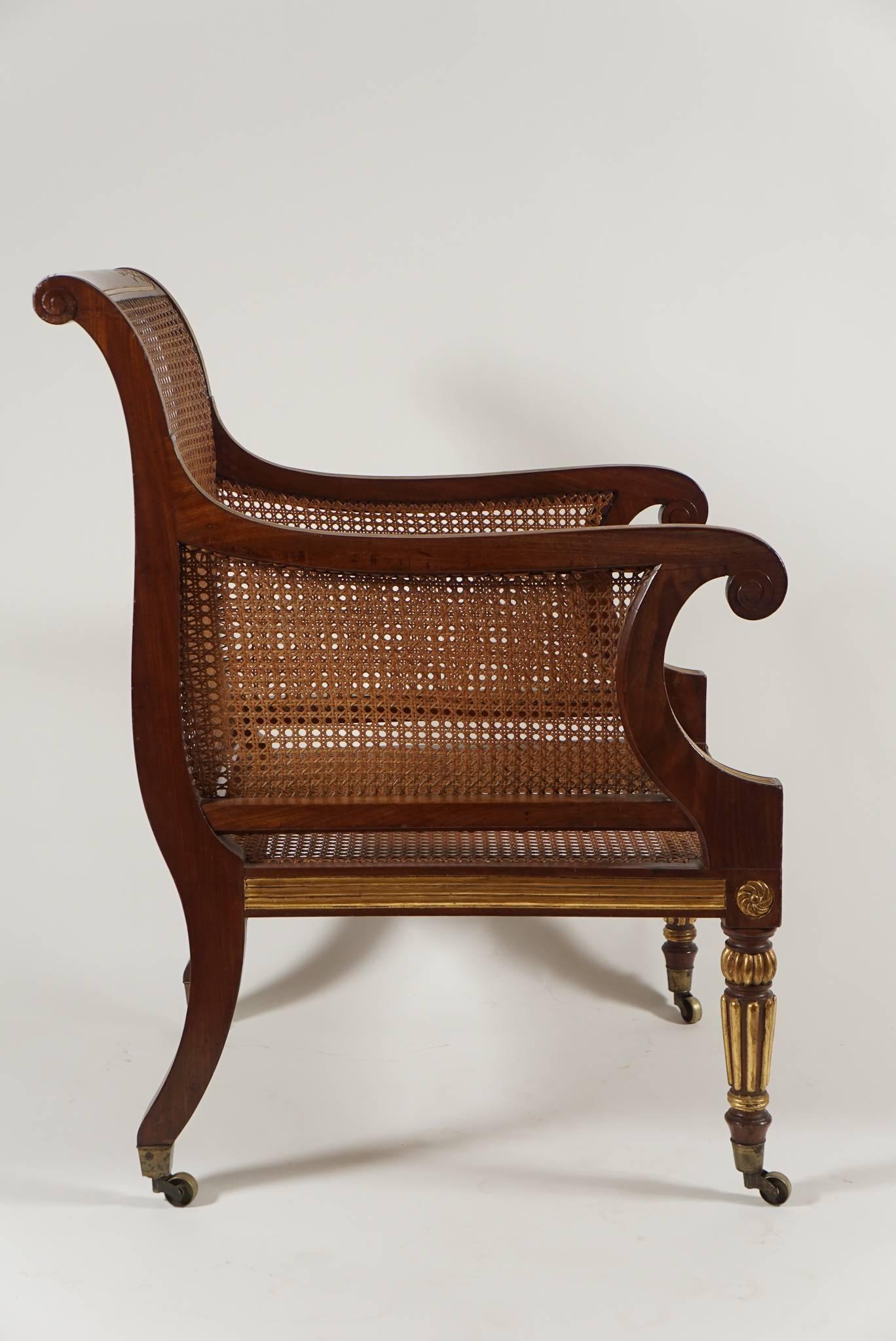 Fine English Regency period mahogany parcel-gilt bergere armchair having caned back, side panels, and seat. Backrest rail having carved and gilt bellflower panel detail with fluted gilt arms terminating in carved volute scrolls and gilt horizontal