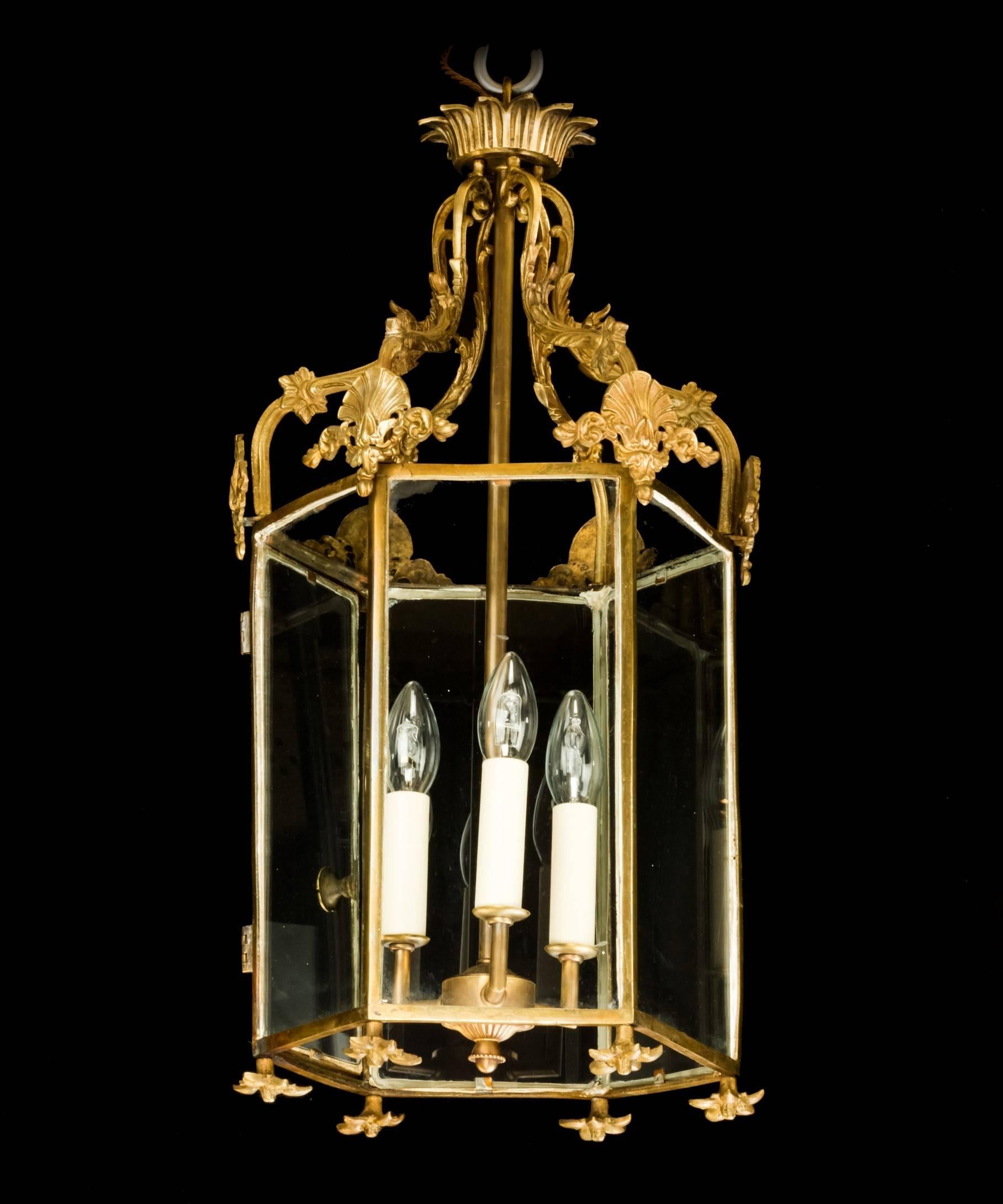 A Georgian style brass hanging lantern in the neoclassical taste; the lantern's hexagonal frame with a crown composed of scrolling acanthus leaves with anthemion’s to the corners of the frame. The lantern's three-light central pendant is modern.