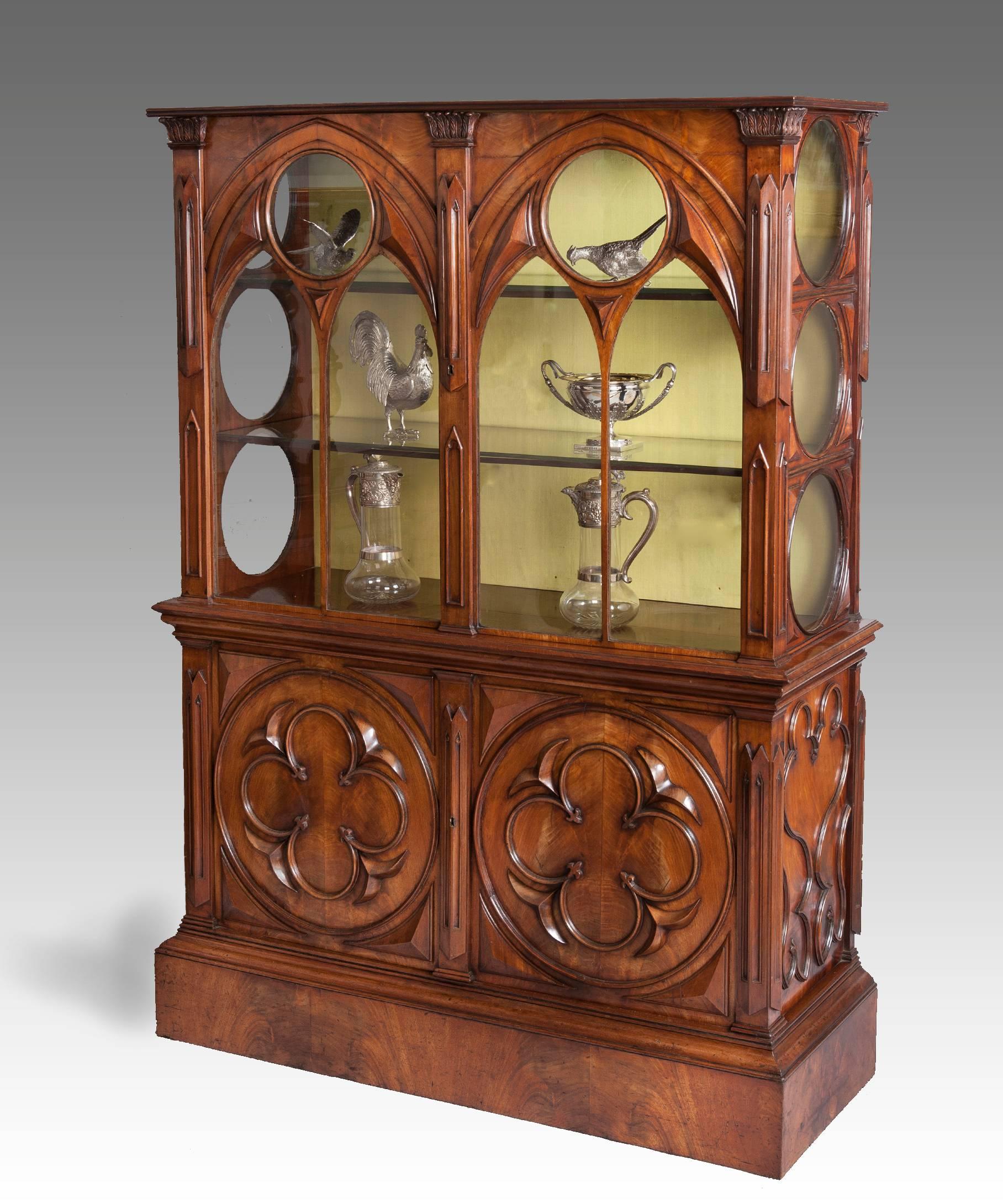 An unusual early 19th century mahogany display cabinet in the Gothic taste; the display cabinet's glazed Gothic arched doors opening to reveal display shelves above a pair of cupboard doors decorated with boldy carved quatrefoils and raised on a