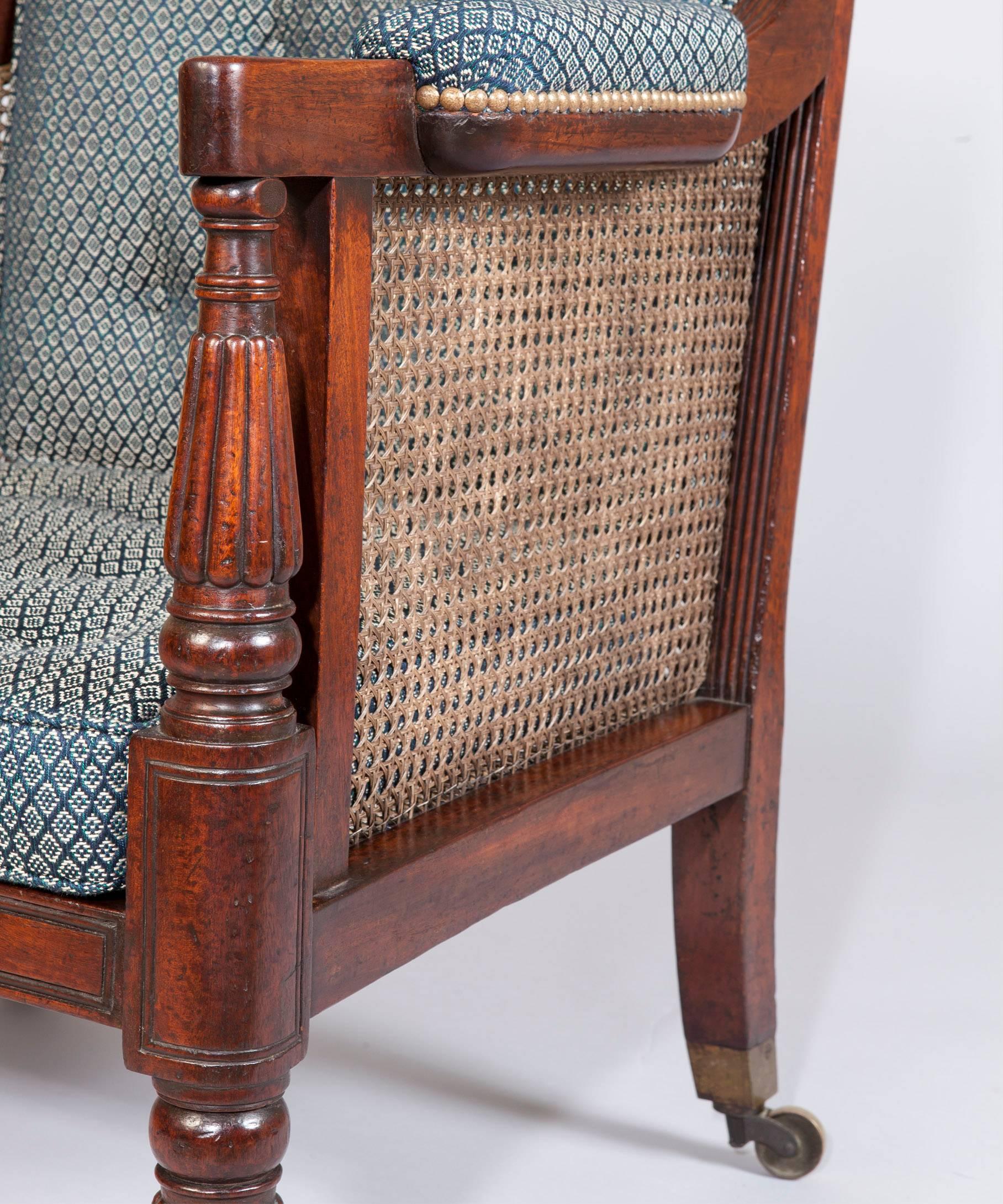 An elegant Regency period mahogany bergere library armchair; the mahogany showwood frame with reeded downswpt arms raised on turned and reeded supports and standing on turned and reeded legs which terminate in the original brass castors; the