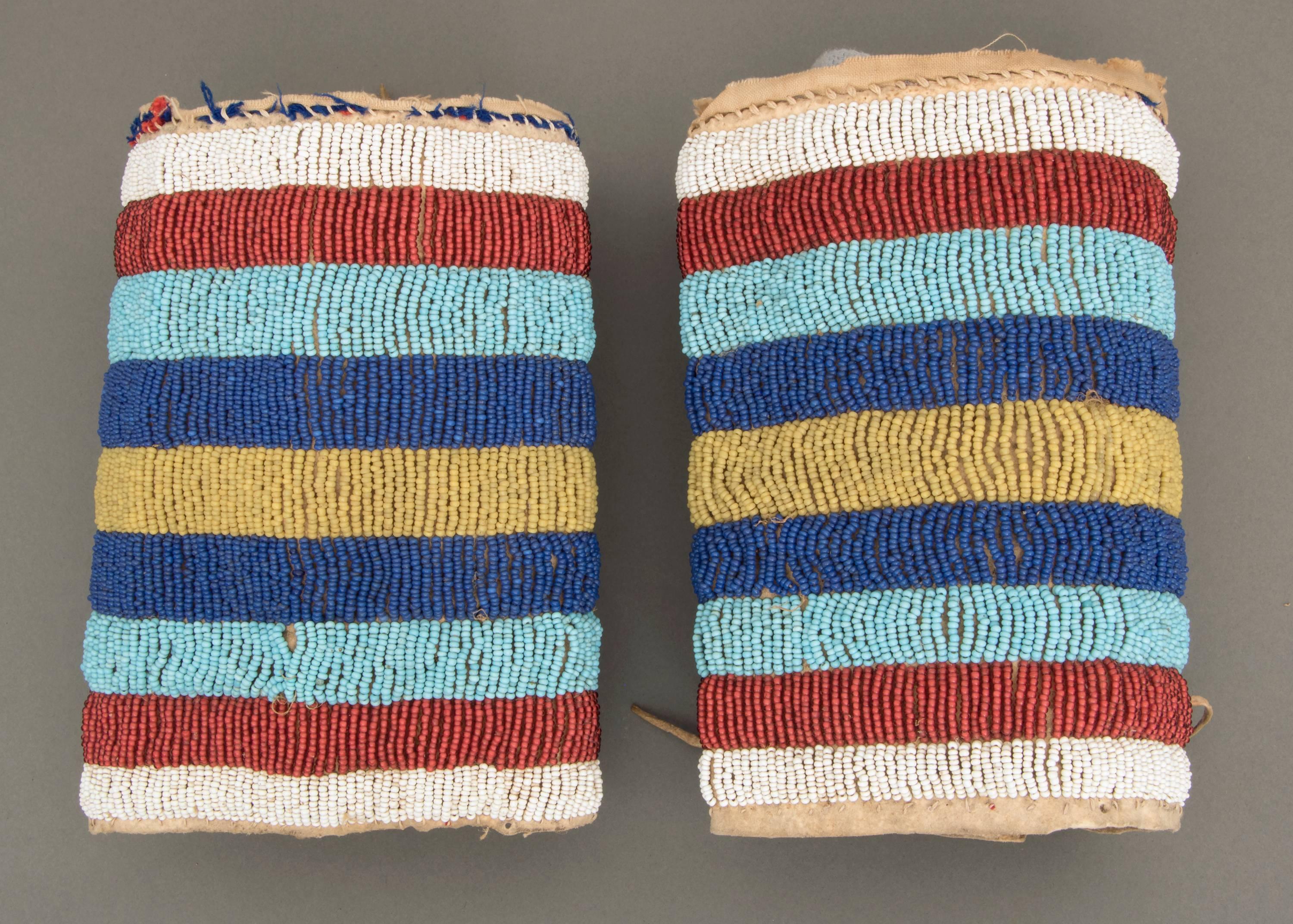 A pair of crow (Plains Indian) beaded ankle cuffs constructed of native tanned hide with glass trade beads in a striped pattern of white, light and dark blue, greasy yellow and white-heart red.
 