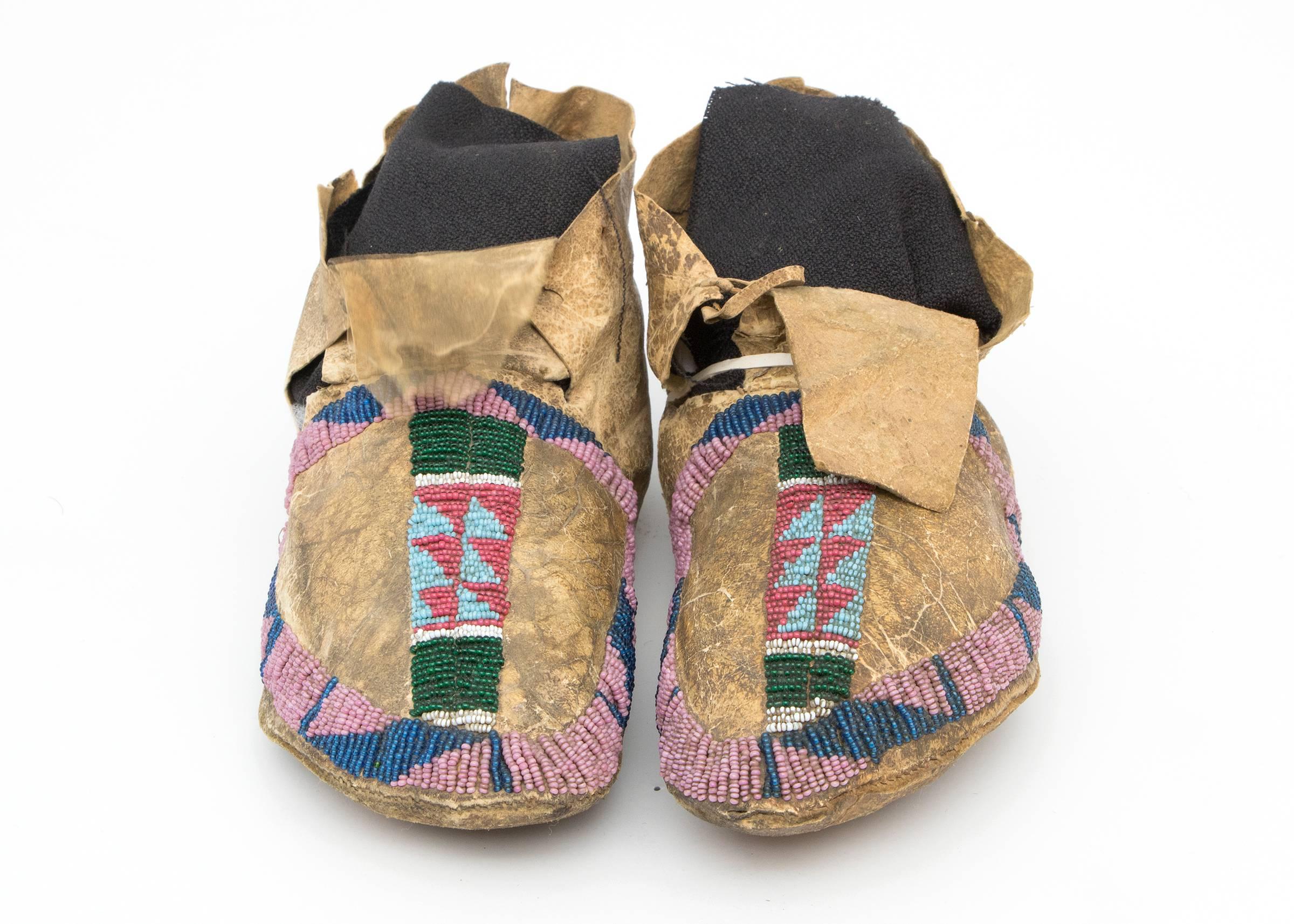 A pair of late Classic period moccasins made for a youth or child. Constructed of native tanned hide and partially beaded around the vamps creating buffalo track motifs in the negative area. Geometric patterns of blue, pink, red and green trade
