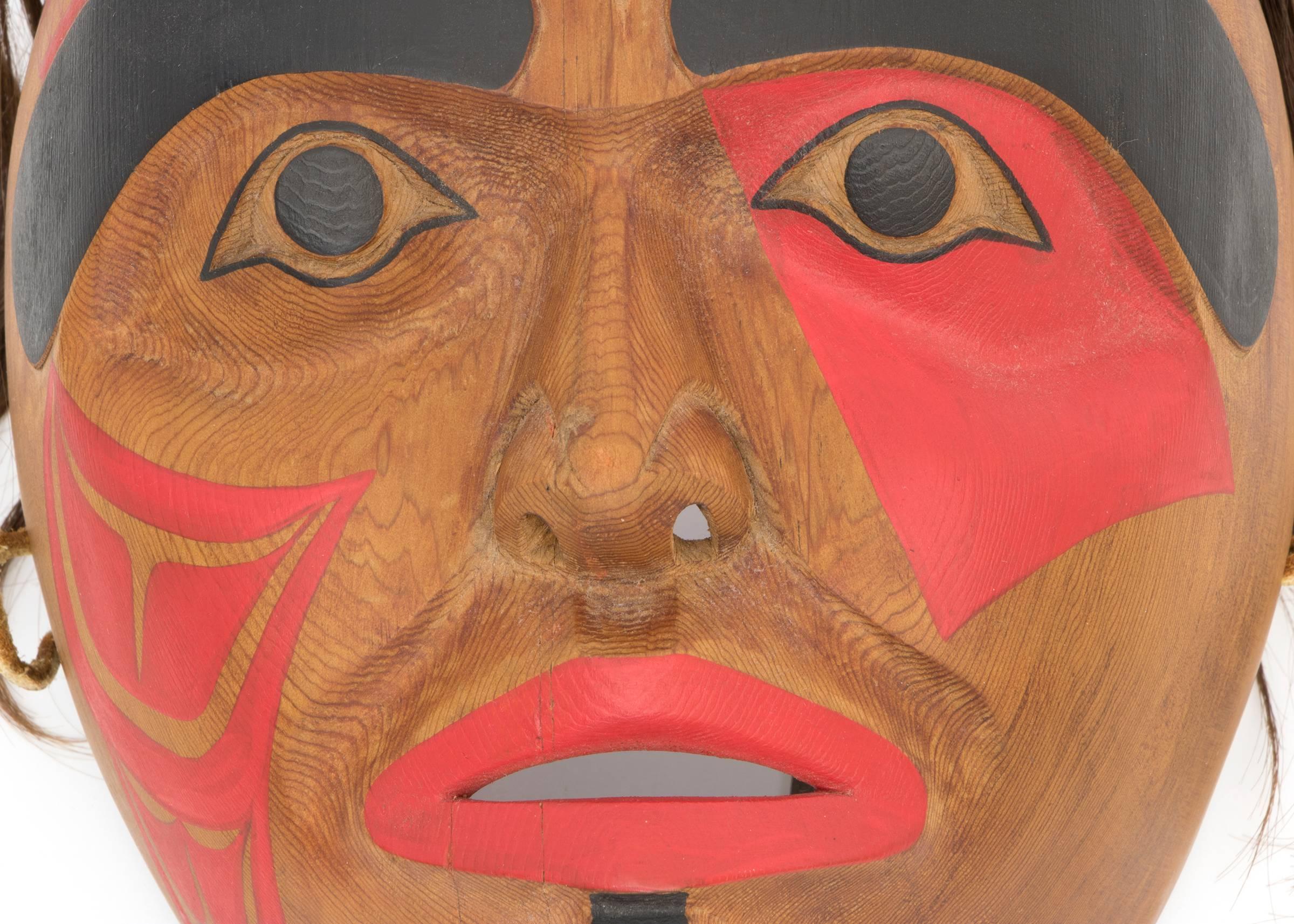 A carved wooden mask originating in the Pacific Northwest Coast. The design on the face is a Native American Killer Whale or Orca motif in black and red. The title, date and the artist's initials, 