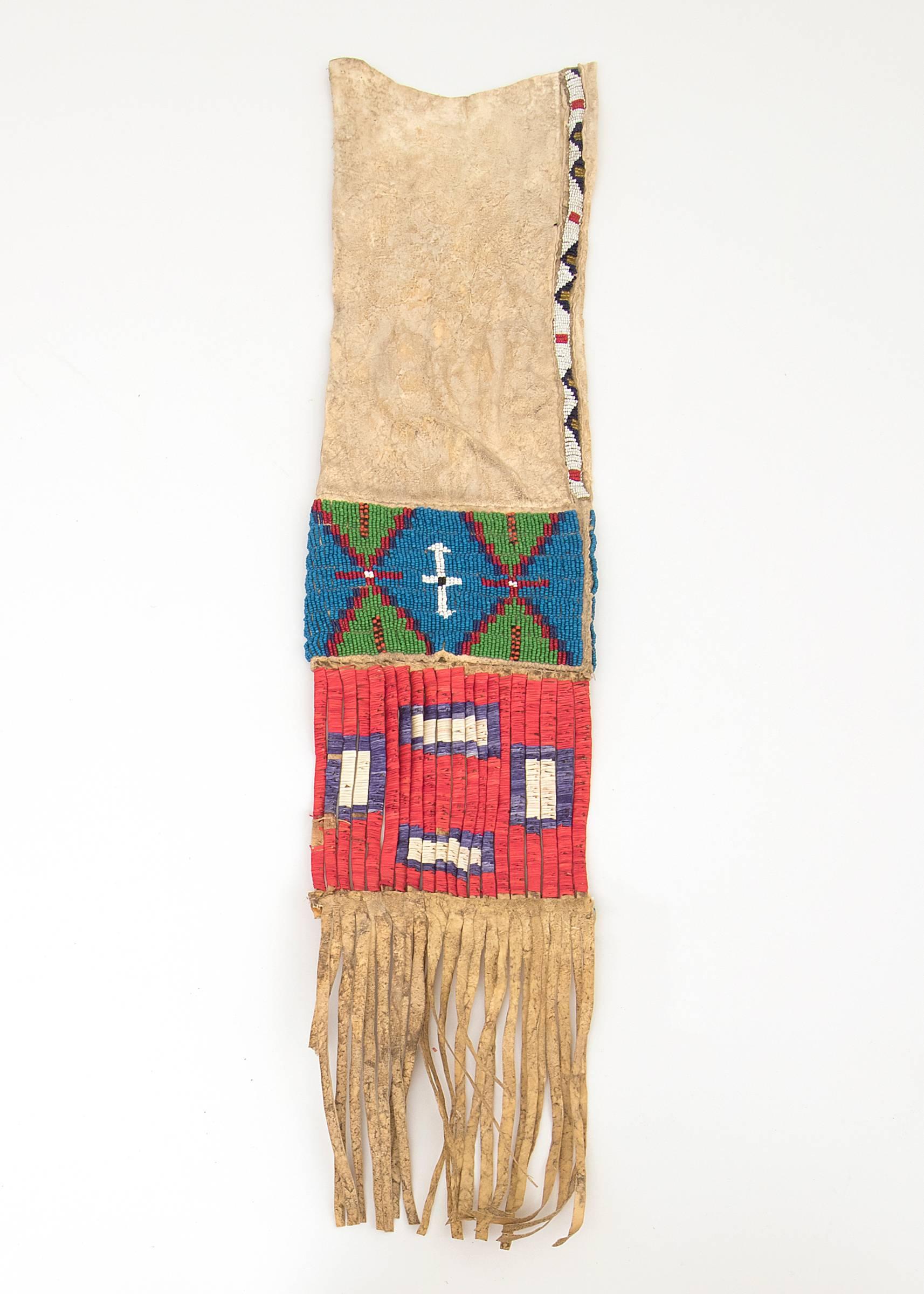 A pipe bag (Tobacco Bag), constructed of native tanned hide and and beaded with stylized tepee motifs in red, blue, orange, green, black and white trade beads. The opposite side has a beaded panel with geometric devices in similar colors. The fringe