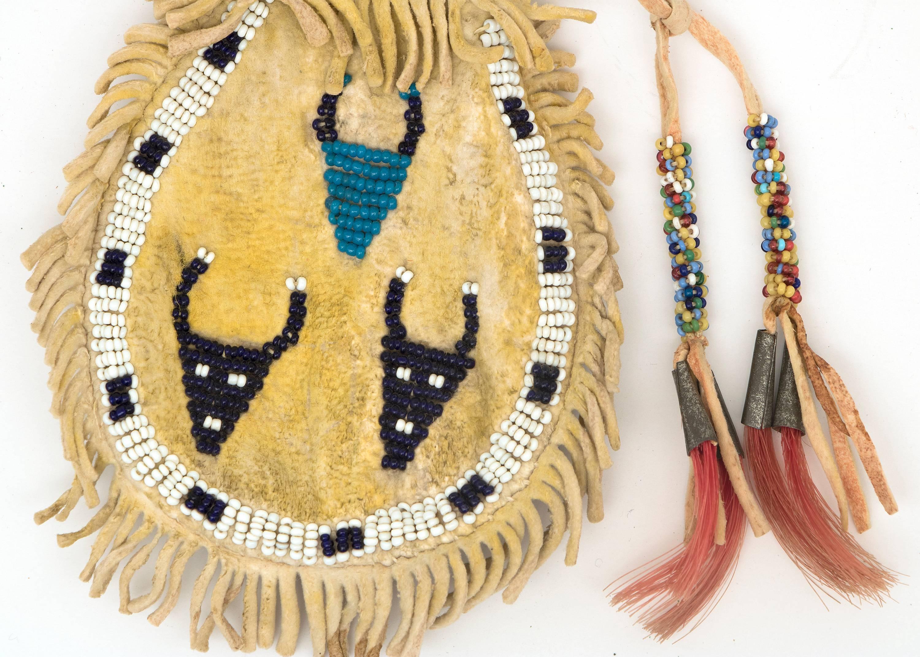 Transitional plains pouch from the last quarter of the 19th century. Constructed of native tanned hide and partially beaded with trade bead. Side 1: three buffalo pictographs; side 2: cross motif; with yellow ochre pigment on both sides and tin-cone