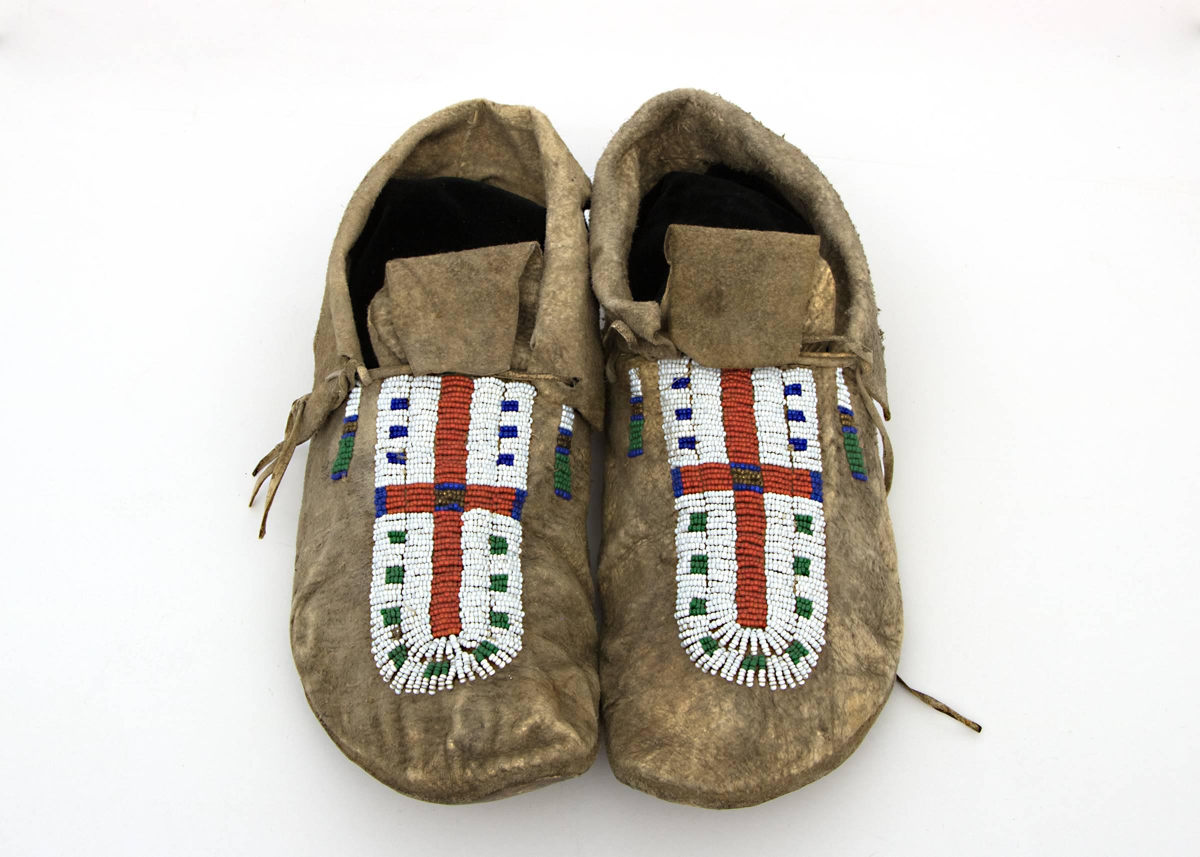 Constructed of native tanned hide with trade beads. Each moc is partially beaded on the vamp with a Red Cross. The cuffs are fringed. 

The Arapaho, members of the Plains Indian culture group, were nomadic with territory extending across parts of