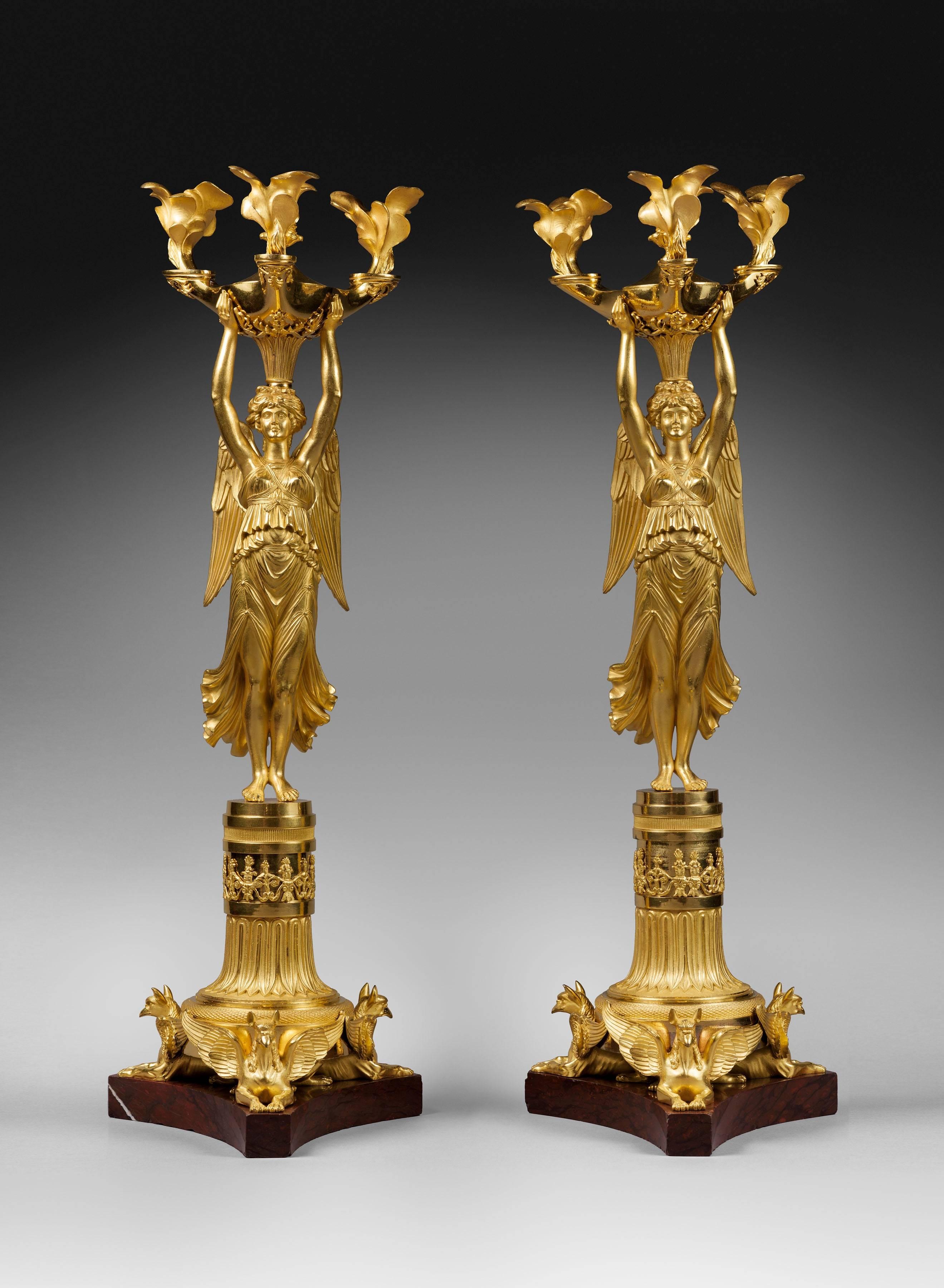 A very fine pair of Empire gilt and patinated bronze three-light candelabra “à la Victoire”, the stem of each, formed as a classically-robed winged Victory whose uplifted arms support a lamp issuing from a pilaster placed upon her head and