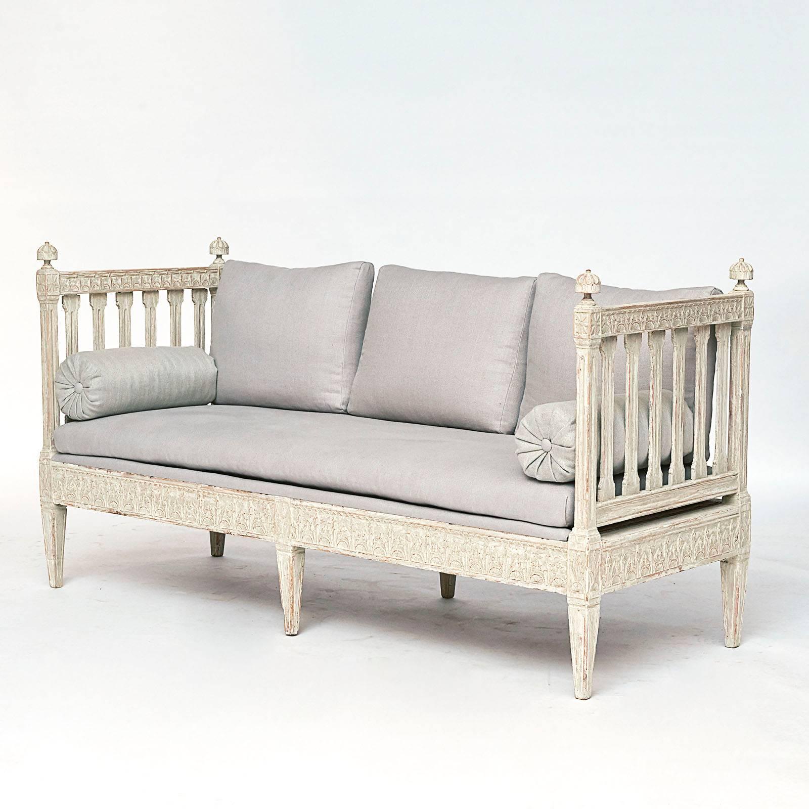 A Swedish late Gustavian painted bench, late 18th-early 19th century, the loose cushion seat and back between slat form sides with leaf carvings and headed with turned finials, a leaf carved frieze raised on square tapered legs. Re-upholstered in