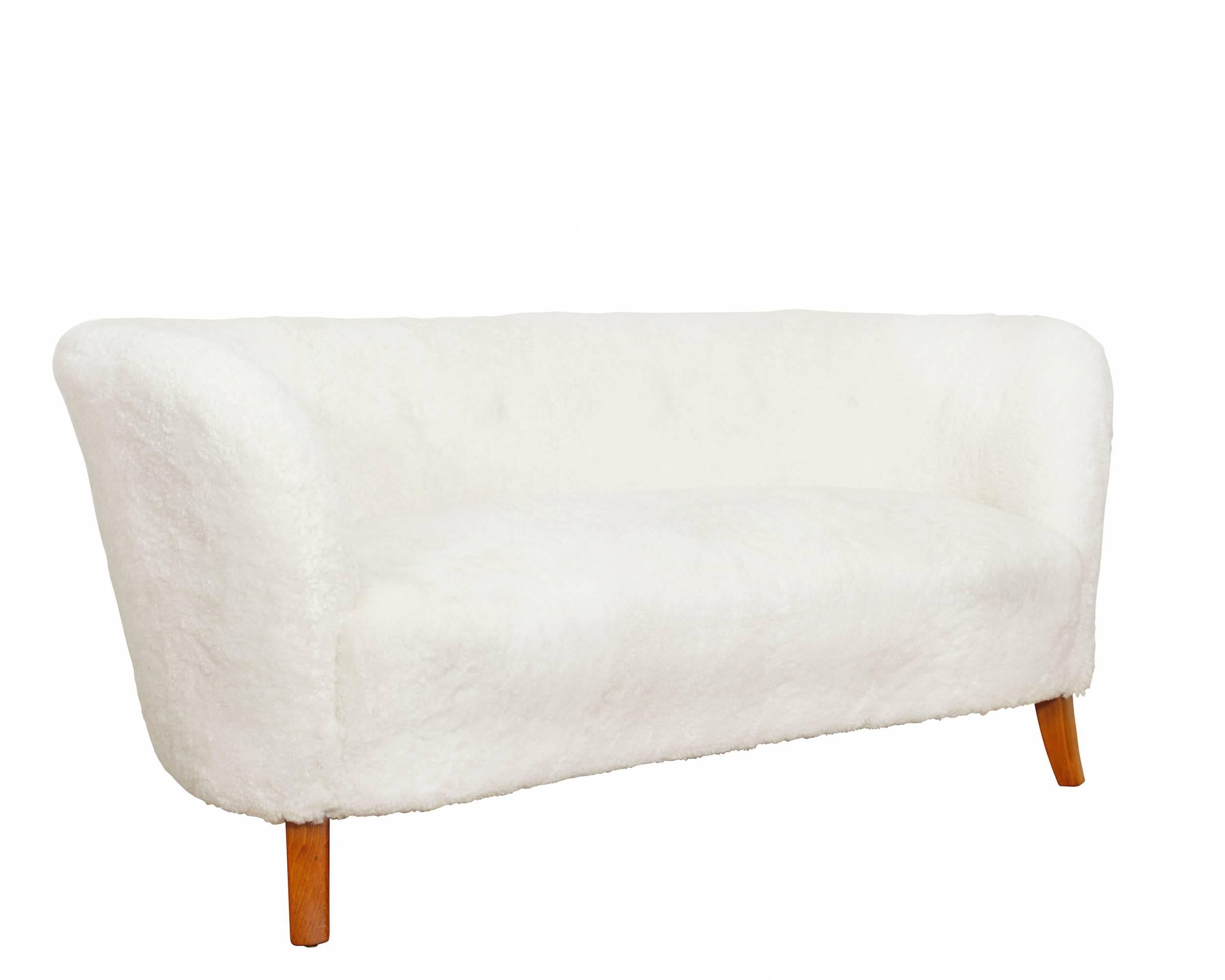 A three-seat curved sofa, upholstered in white sheepskin, raised on stained beech legs. 
Manufactured by Georg Kofoed.