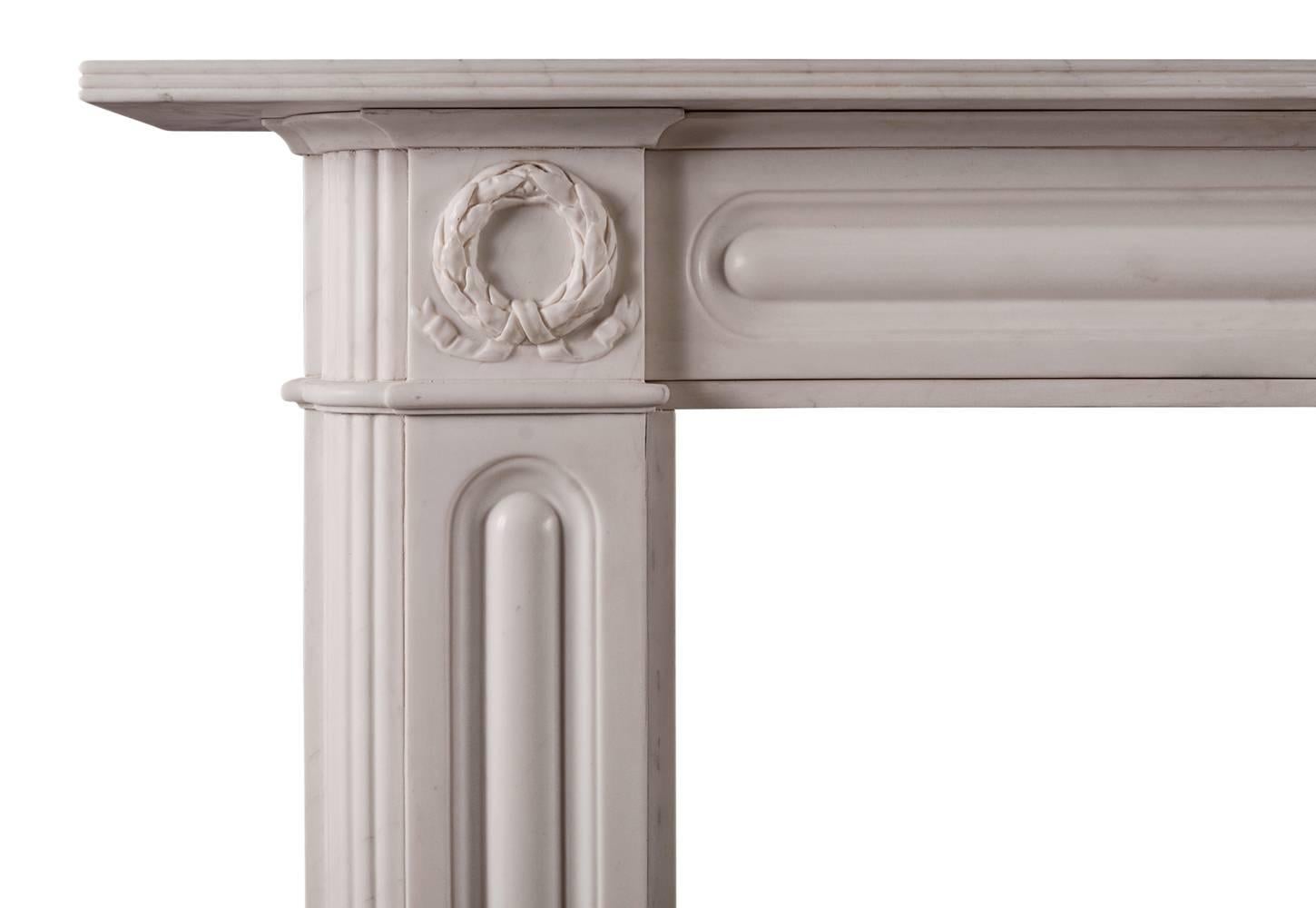 Une cheminée anglaise en marbre blanc de style Regency. Les jambages à panneaux sont ornés d'une moulure en arc sur les retours et d'une couronne sculptée avec des rubans noués. La frise lambrissée est surmontée d'une tablette cannelée. Une copie de