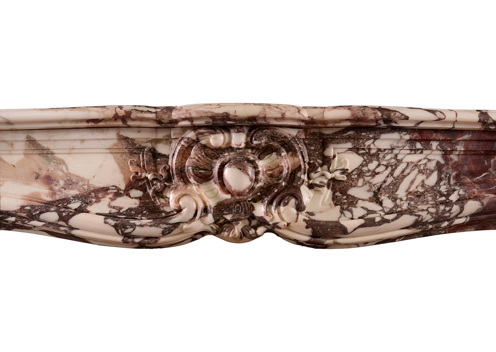 Cheminée Louis XV française du XVIIIe siècle en marbre Violette. Les jambages à volutes sont surmontés de coquilles sculptées, la frise lambrissée est ornée d'un cartouche central. Au-dessus, une étagère façonnée et moulurée. Une pièce